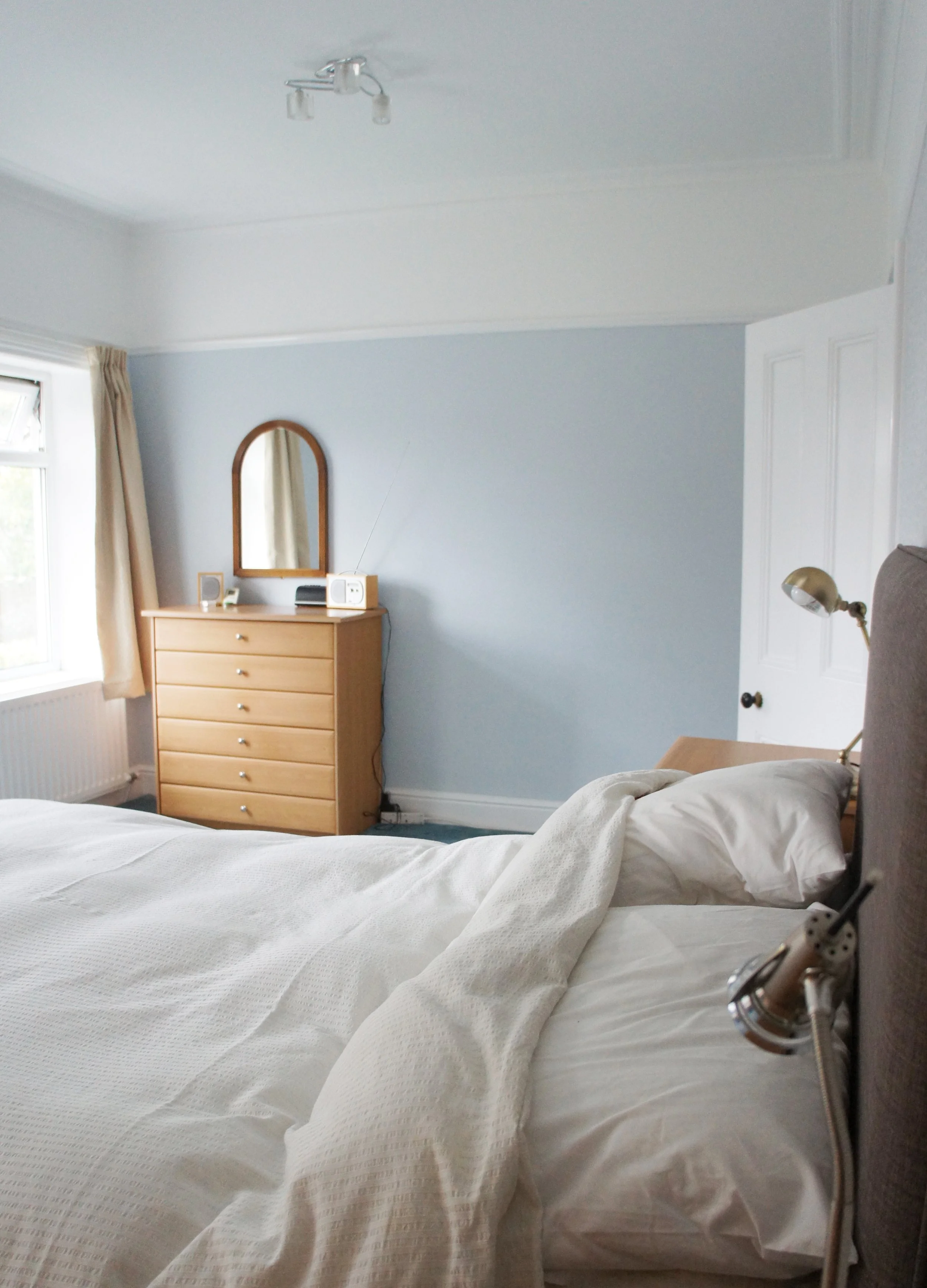 A bedroom with pale blue painted walls, wooden chest of drawers and white door and trim