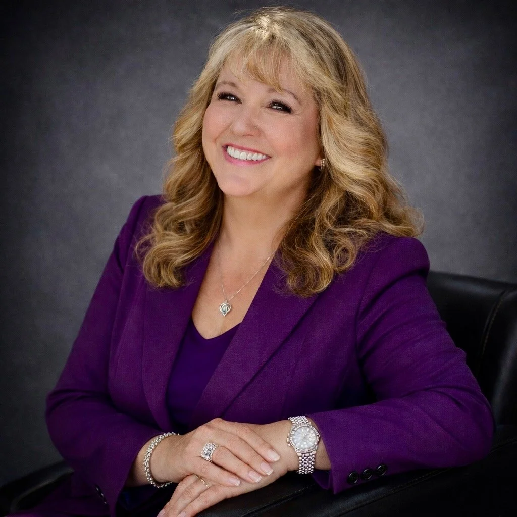 A woman with blonde curly hair, smiling outdoors near a tree, wearing a white lace top and a silver necklace with a heart-shaped pendant.