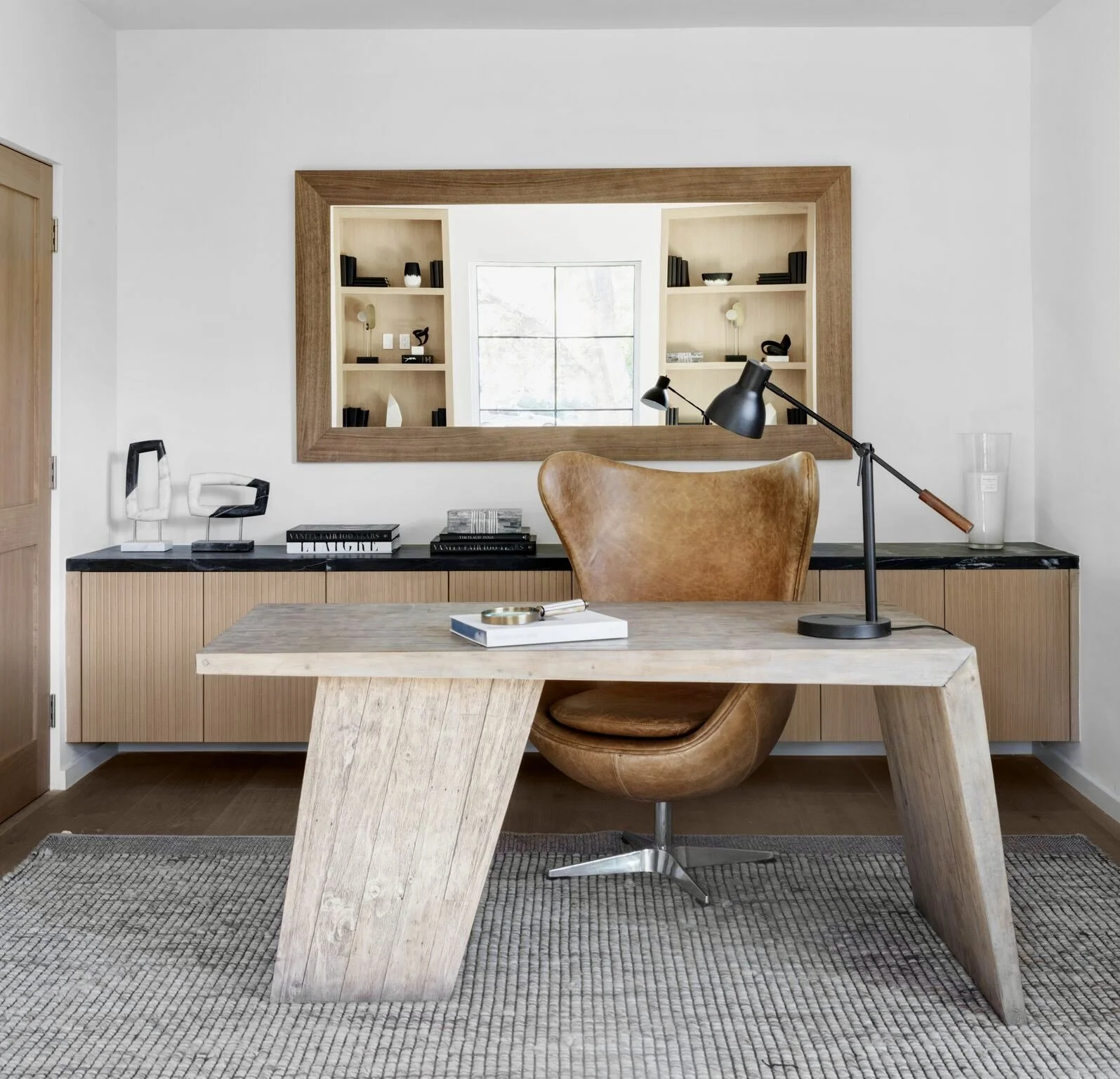 Modern home office with wooden desk, leather swivel chair, black desk lamp, large mirror, dining sideboard, decorative objects, and window reflection.  Rachael Goddard Design Studio Calabasas