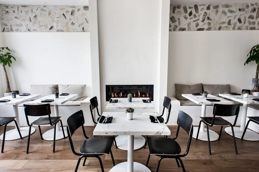 Rachael Goddard Design Studio Calabasas Elegant restaurant interior with white tables, black chairs, and a modern fireplace. Decor includes potted plants, stone accents on the walls, and neatly set tables with black napkins and small plants.