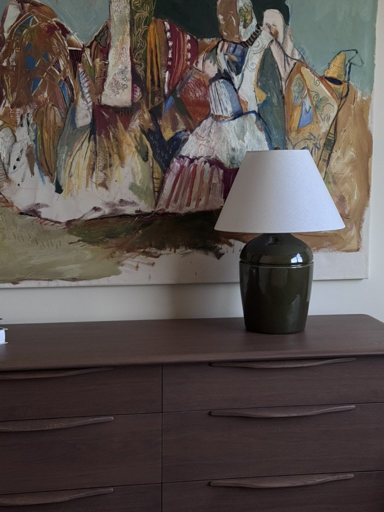 Rachael Goddard Design Studio Calabasas A dark green ceramic table lamp with a white lampshade on a wooden dresser in a room with abstract colorful art on the wall.