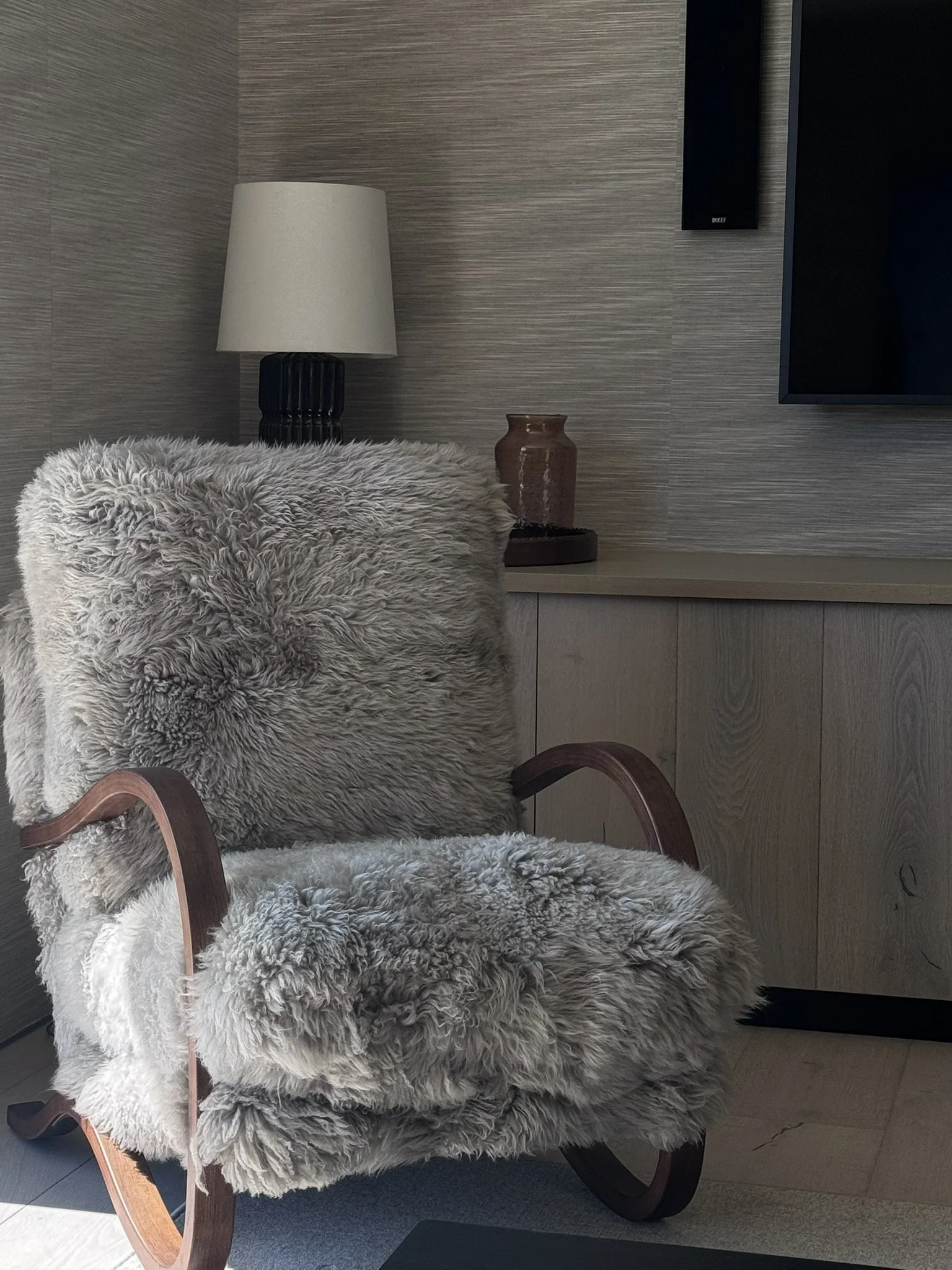 Rachael Goddard Design Studio Calabasas A cozy rocking chair with a wooden frame and fluffy beige fur upholstery and cushions, placed in front of a beige cabinet with a lamp and decorative jar on top, with a textured wall
