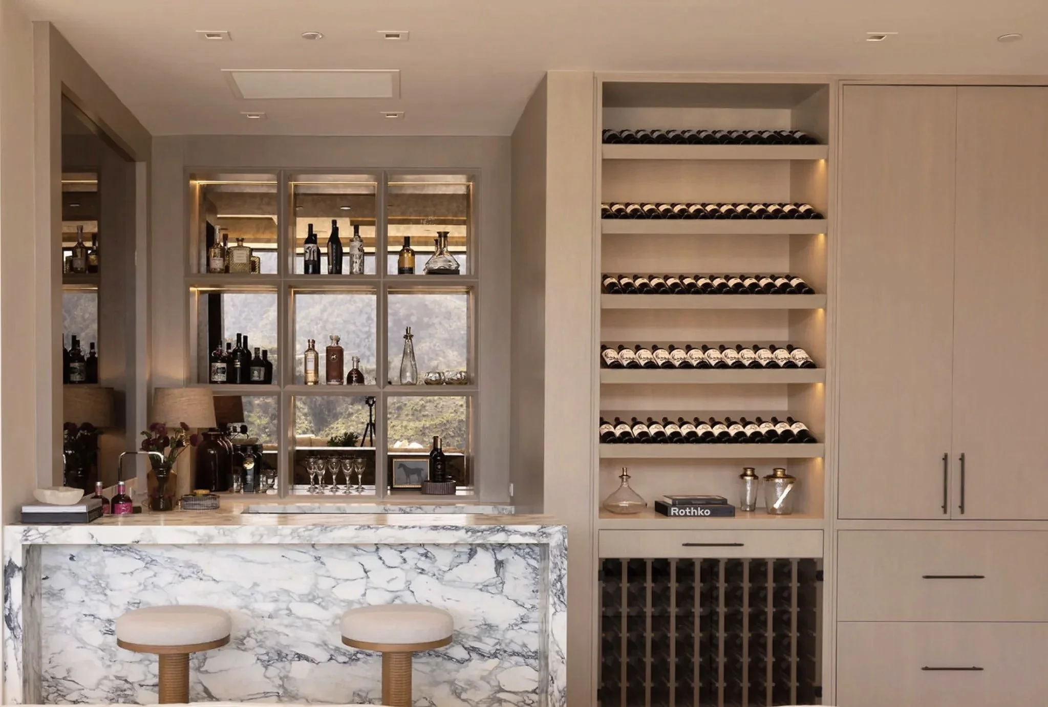 Rachael Goddard Design Studio Calabasas Modern home bar with a marble countertop, bar stools, and built-in shelves stocked with wine bottles and glassware. Decor includes bottles, vases, and decorative items.