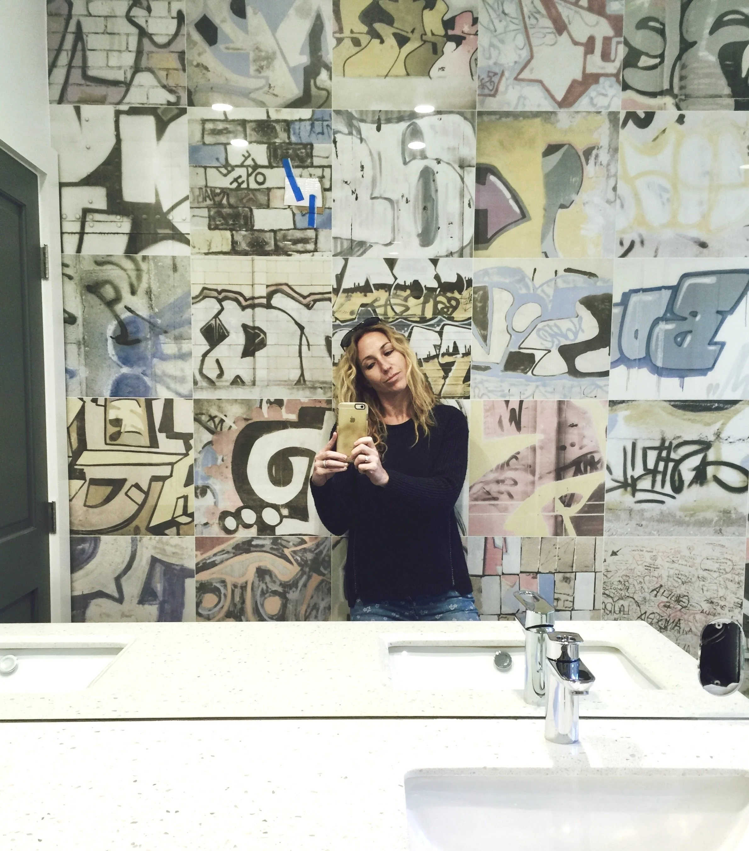 A woman taking a mirror selfie in a bathroom with graffiti-style artwork on the tiled wall behind her.  Rachael Goddard Design Studio Calabasas
