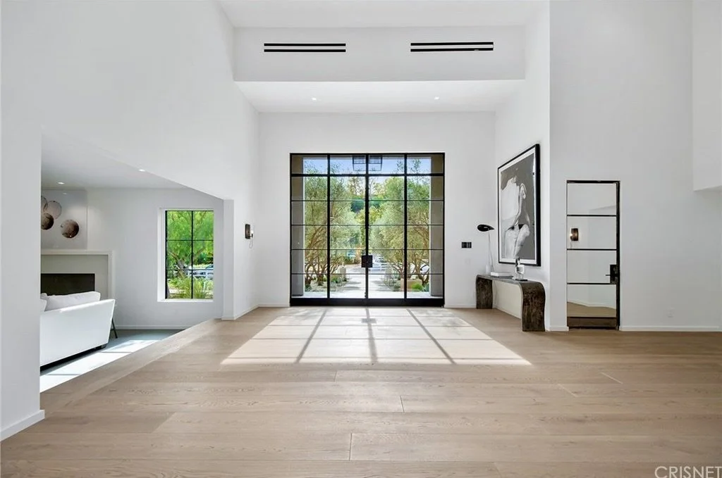 Interior view of a modern, minimalist living room with white walls, wooden floors, and large glass doors leading outside, with sunlight casting shadows on the floor.  Rachael Goddard Design Studio Calabasas