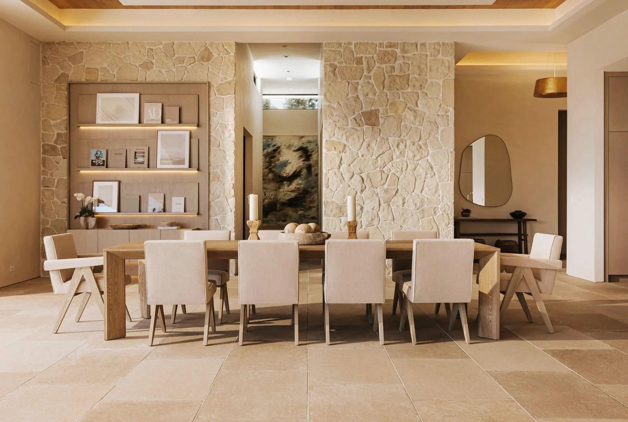 Rachael Goddard Design Studio Calabasas Modern dining room with a large wooden table surrounded by cream-colored chairs, stone accent wall, built-in shelves with artwork and decor, beige tiled floor, and minimalist decor