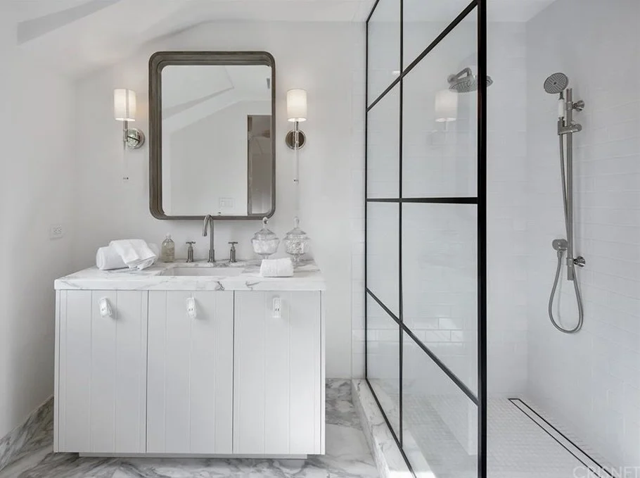 Modern bathroom with white vanity, marble countertop, large mirror, wall-mounted sconces, glass jars, and a walk-in shower with black framing and a handheld showerhead.  Rachael Goddard Design Studio Calabasas