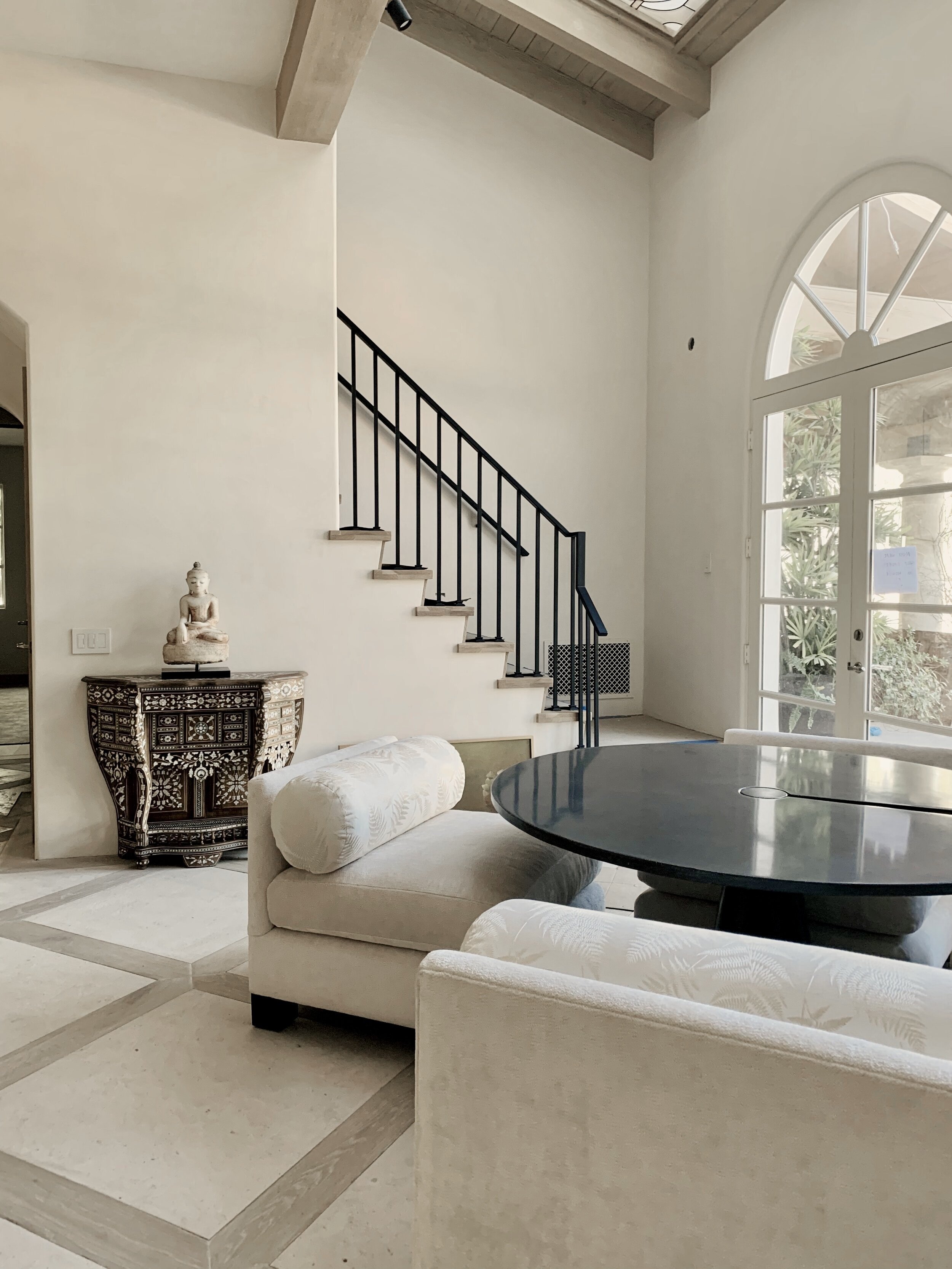 Rachael Goddard Design Studio Malibu Retreat living room with white sofas, black round table, sculptural decor, staircase, and arched window.