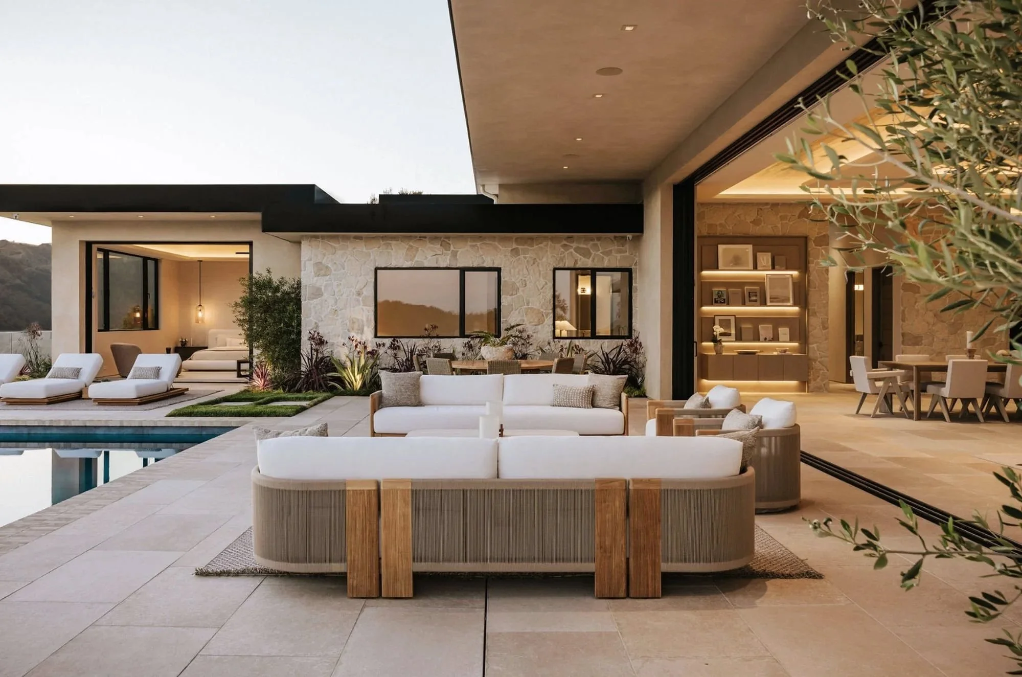Rachael Goddard Design Studio Calabasas Modern outdoor patio with white sofas, lounge chairs, and a pool, adjacent to a contemporary house with a stone wall and indoor living area visible through large open sliding doors.