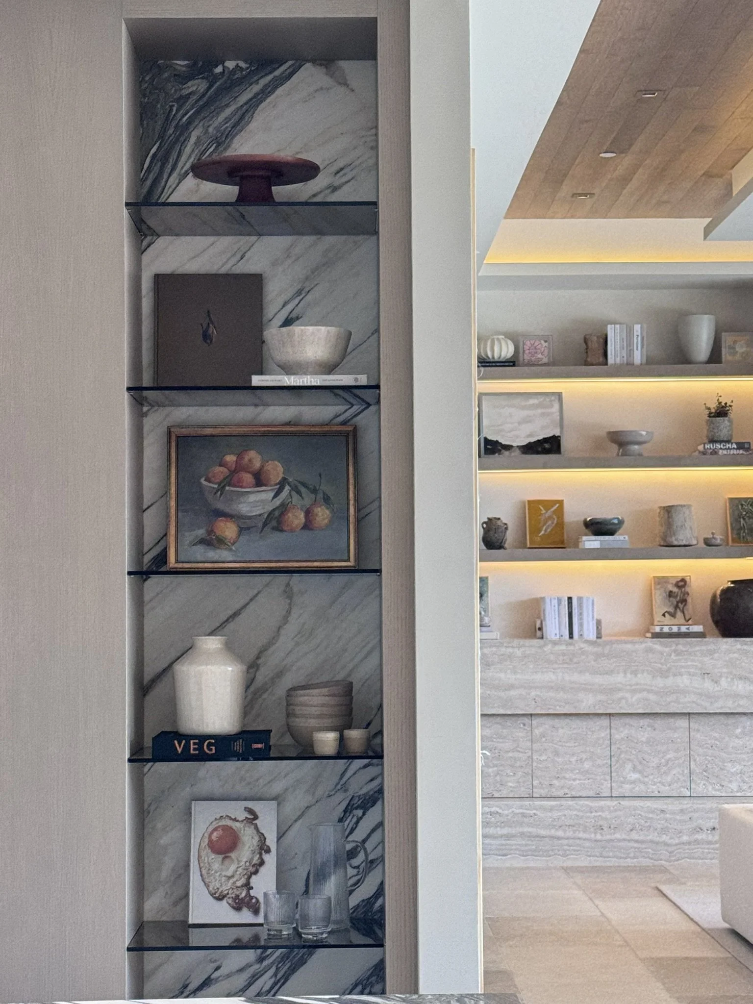 Rachael Goddard Design Studio Calabasas Open shelving with decorative items such as a red cake stand, a beige bowl, a framed artwork of peaches, a large white vase, stacks of bowls, a book titled 'VEG', and smaller objects