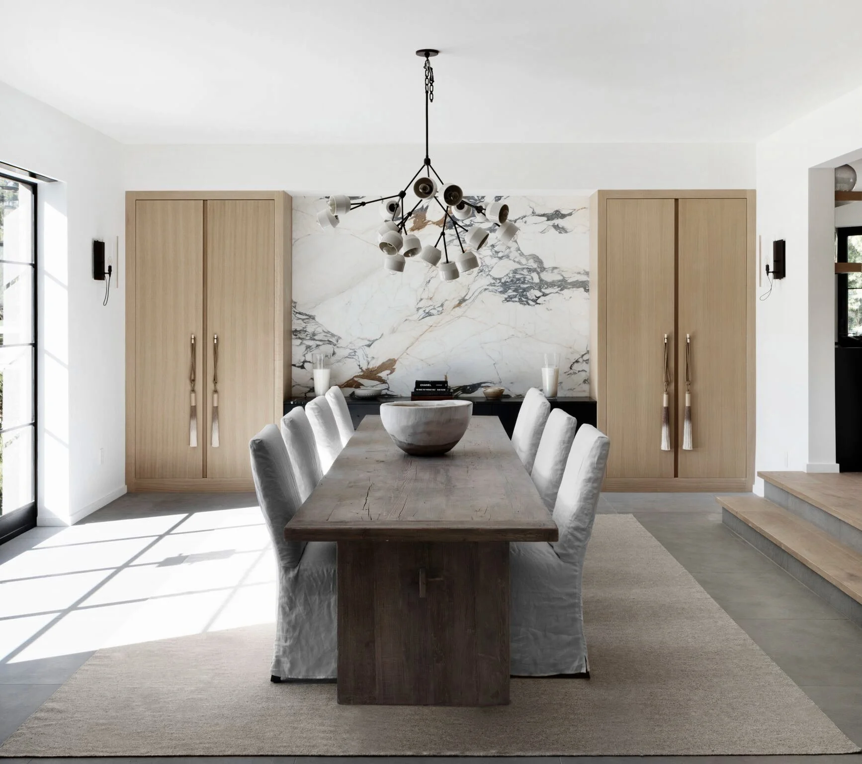 Modern dining room with wooden table, six upholstered chairs, marble backsplash, wooden cabinets, chandelier, and large window  Rachael Goddard Design Studio Calabasas