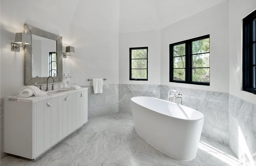 Modern white bathroom with a standalone bathtub, a large mirror, and three black-framed windows  Rachael Goddard Design Studio Calabasas