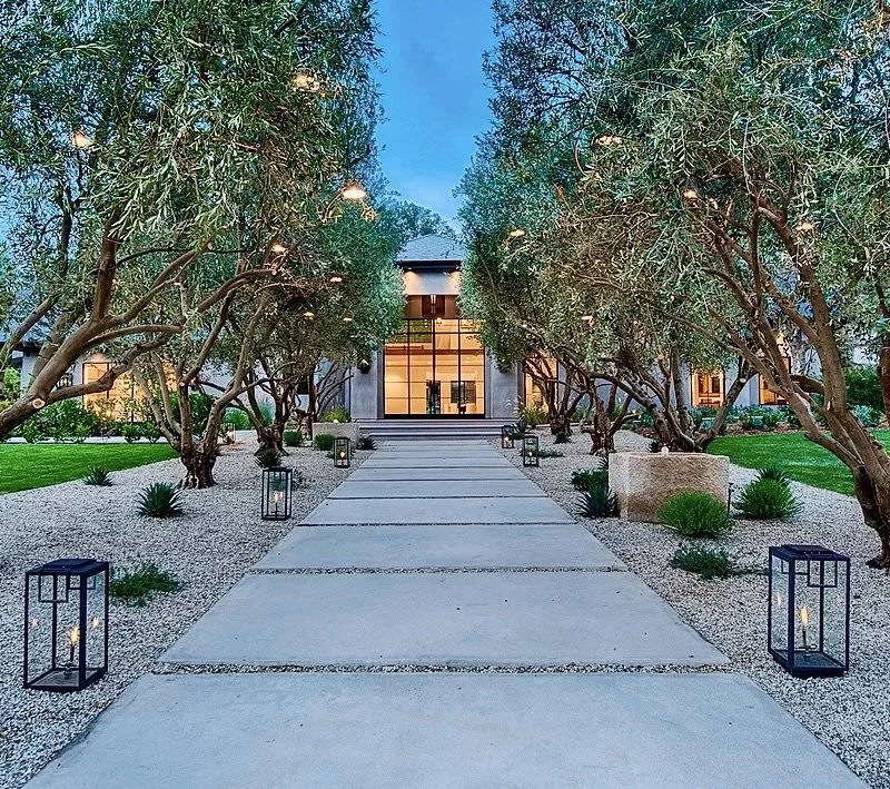 A modern house entrance with a concrete walkway lined with lantern-style lights, mature trees, and well-kept landscaping leading to a glass door.  Rachael Goddard Design Studio Calabasas
