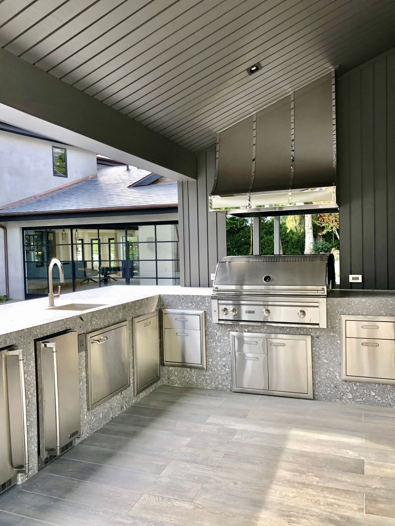 Modern outdoor kitchen with stainless steel appliances, granite countertops, and a view of a house with large windows and trees in the background.  Rachael Goddard Design Studio Calabasas