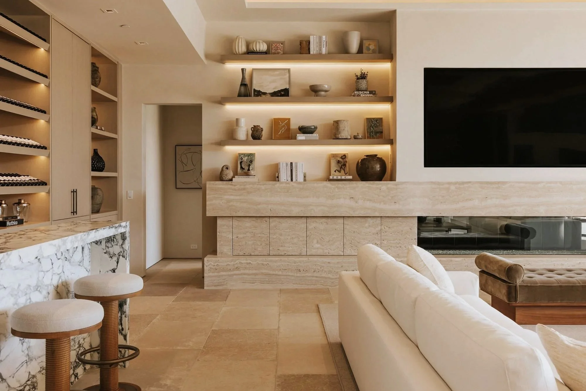 Rachael Goddard Design Studio Calabasas Living room with beige and cream decor, featuring built-in shelves with decorative items, a large white sofa, and a wall-mounted flat-screen TV.