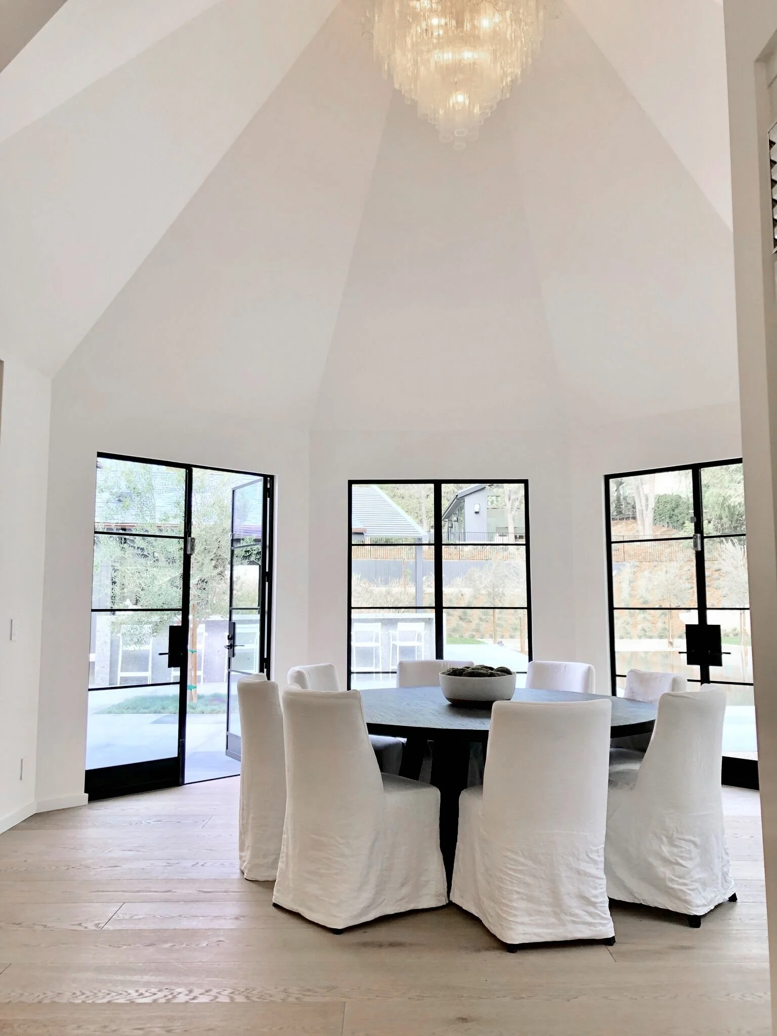 Bright dining room with white walls, a vaulted ceiling, a large chandelier, round black dining table, and nine white slipcovered chairs. Large glass doors and windows surround the space  Rachael Goddard Design Studio Calabasas
