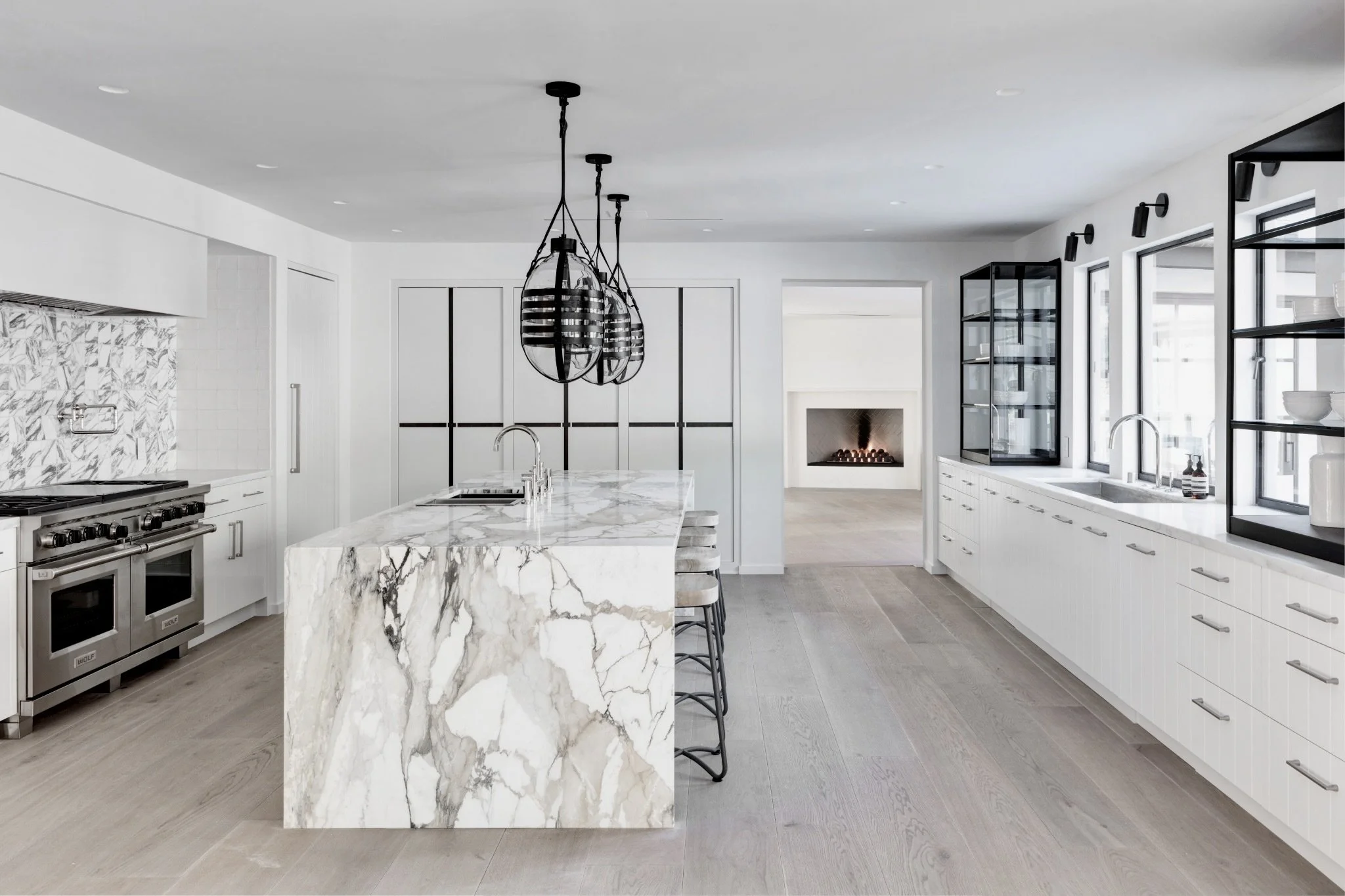 Modern kitchen with white cabinets, marble island, stainless steel appliances, black pendant lights, and a fireplace in the background.  Rachael Goddard Design Studio Calabasas