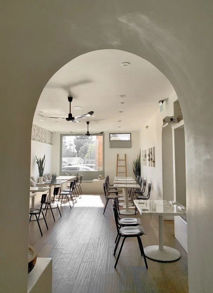 Rachael Goddard Design Studio Calabasas tInterior view of a bright, cozy cafe with white walls, marble tables, black chairs, and large windows letting in natural light. Decor includes potted plants, wall art,
