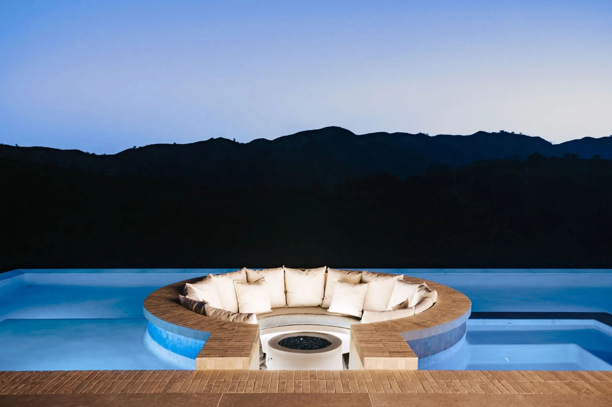 Rachael Goddard Design Studio Calabasas A circular hot tub with embedded seating and beige cushions, surrounded by a deck, set against a backdrop of dark mountains under a clear evening sky.