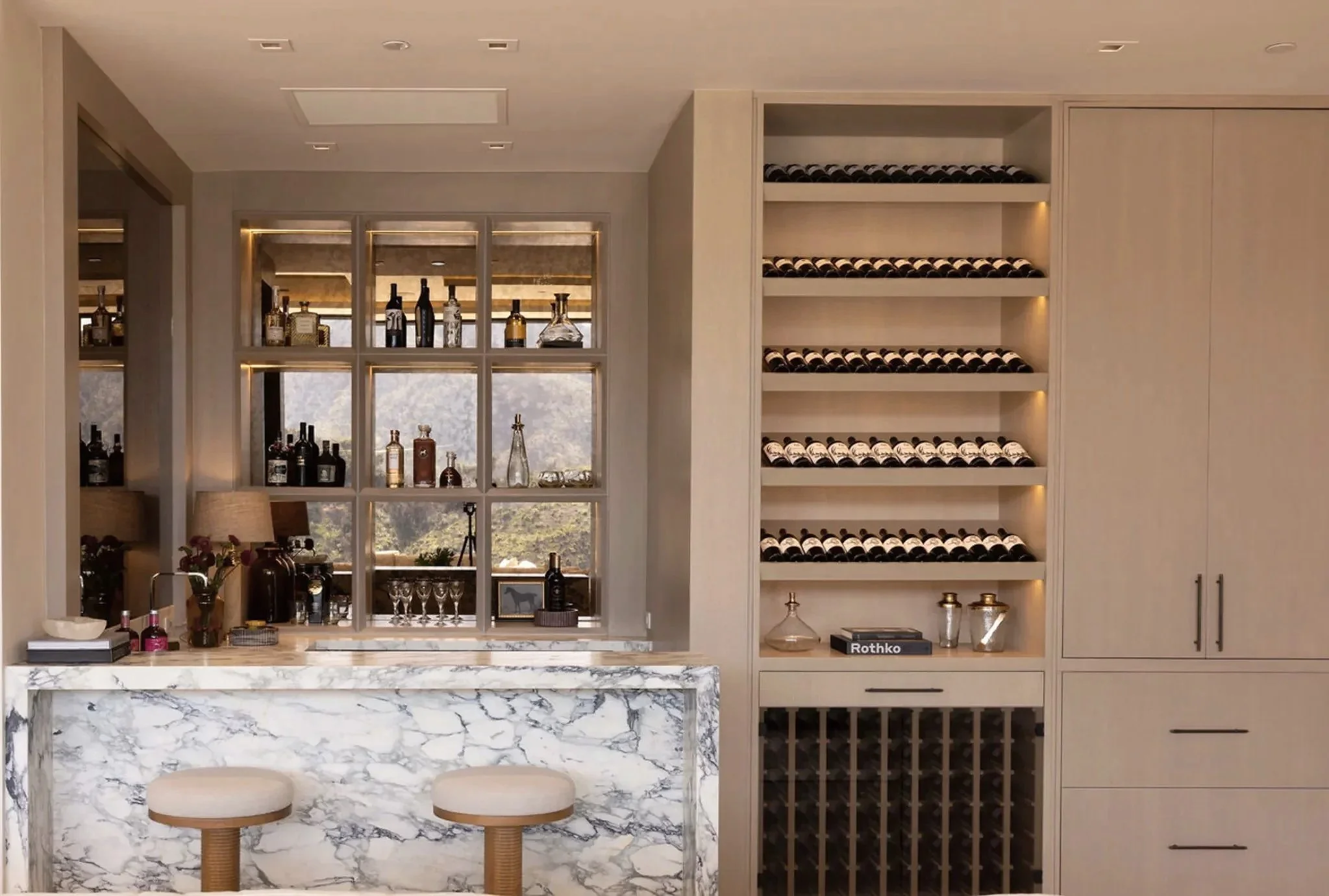 Rachael Goddard Design Studio Calabasas Modern bar area with marble countertop and two bar stools, behind a glass shelving unit with bottles and glassware, adjacent to a wall with wine bottles stored on shelves and a wine rack below.