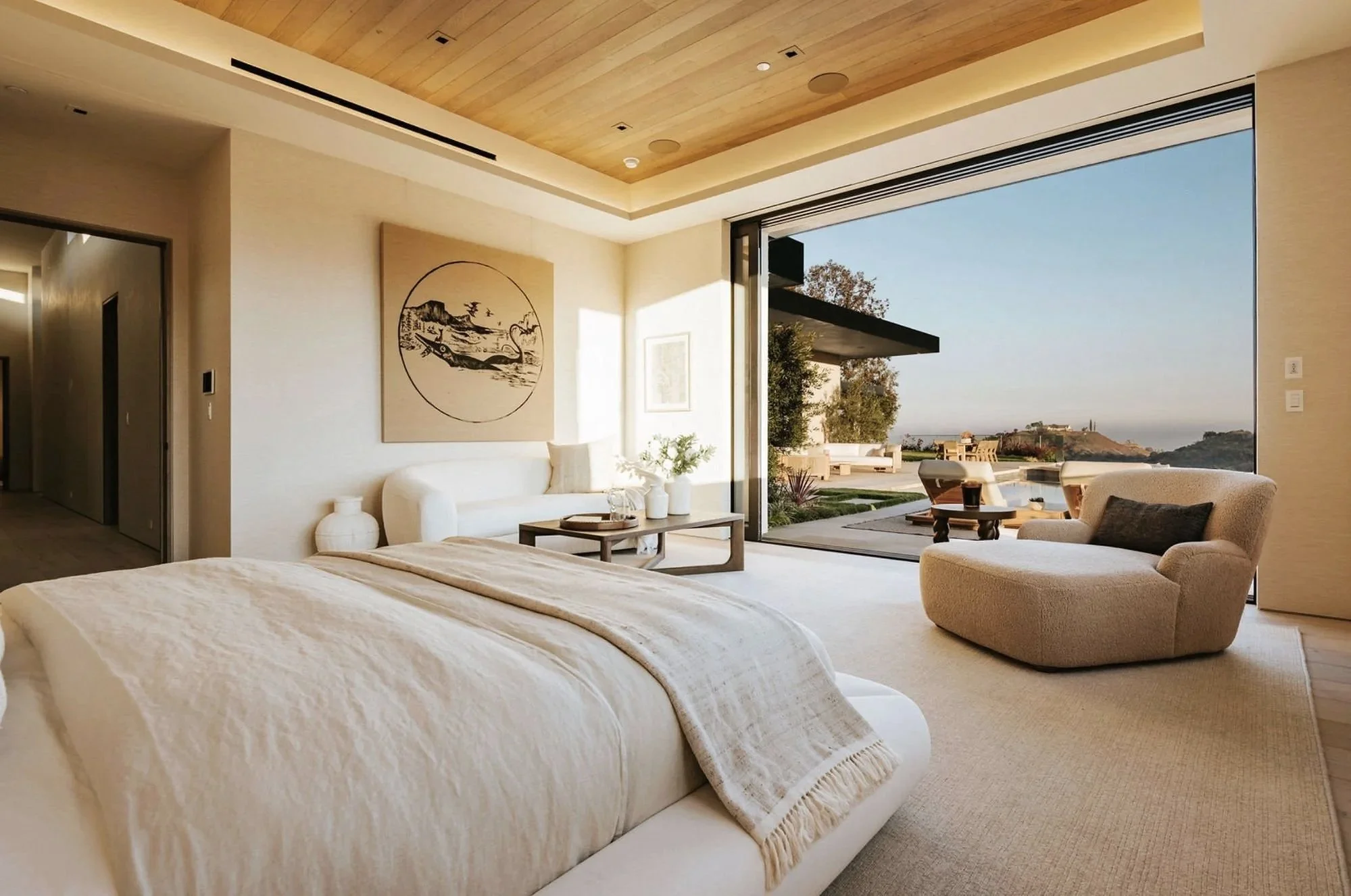 Rachael Goddard Design Studio Calabasas Modern bedroom with large glass sliding door leading to outdoor patio overlooking hills and ocean, featuring neutral-colored furniture and minimalist decor.