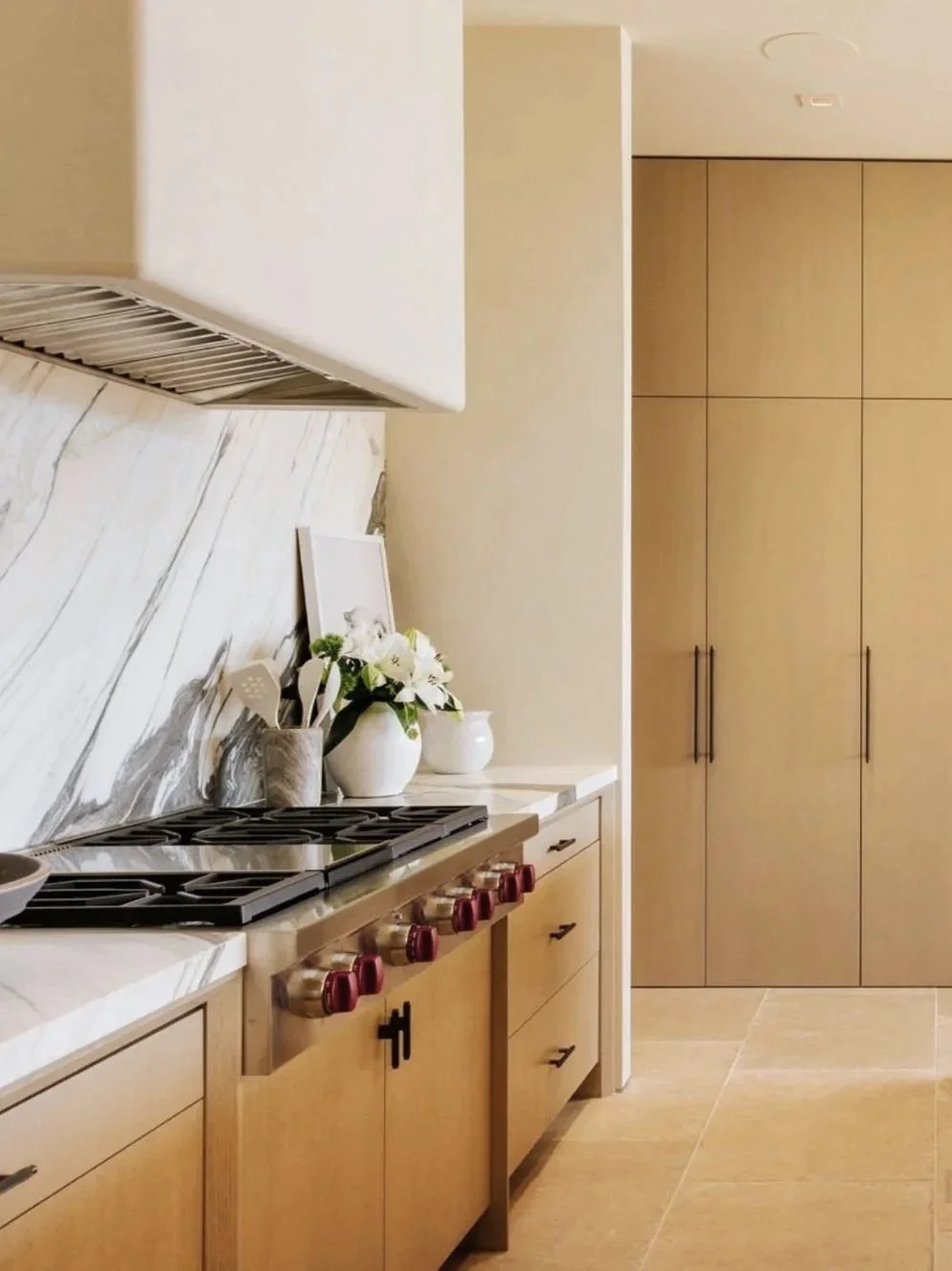 Rachael Goddard Design Studio Calabasas Modern kitchen with beige cabinetry, a gas stove with maroon knobs, a white marble backsplash, and a countertop decorated with white flowers and small white jars.