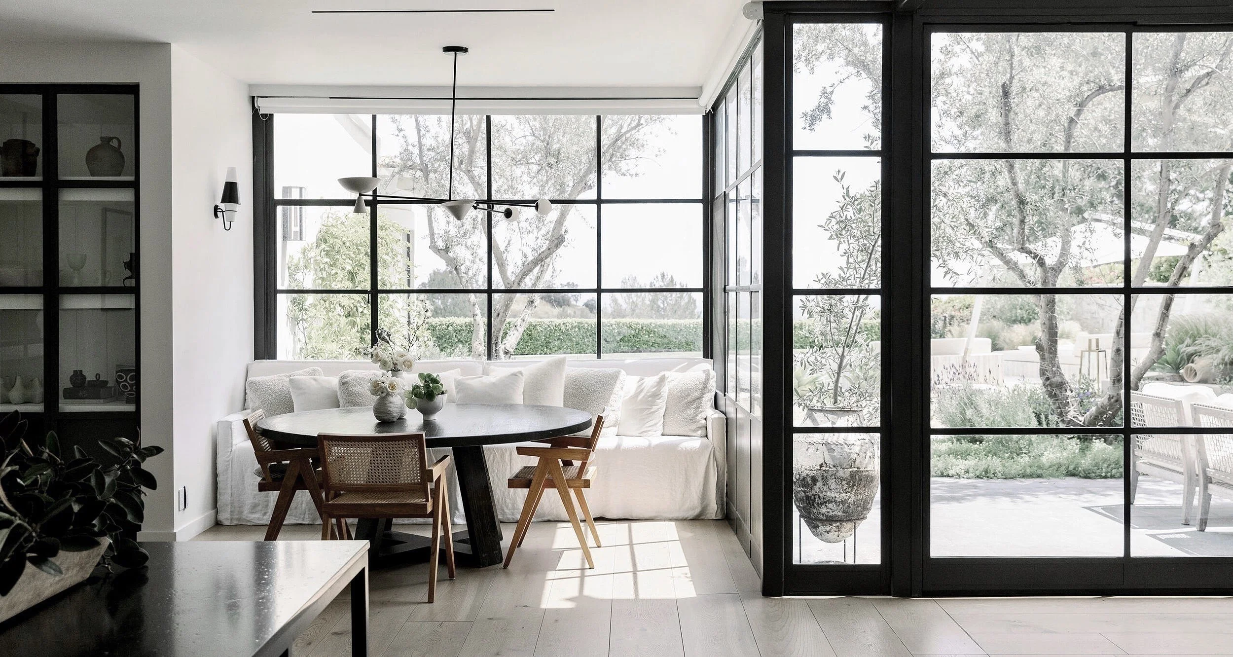 Bright interior living space with large windows, a white sofa, a round table with chairs, and outdoor patio visible through glass doors.  Rachael Goddard Design Studio Calabasas