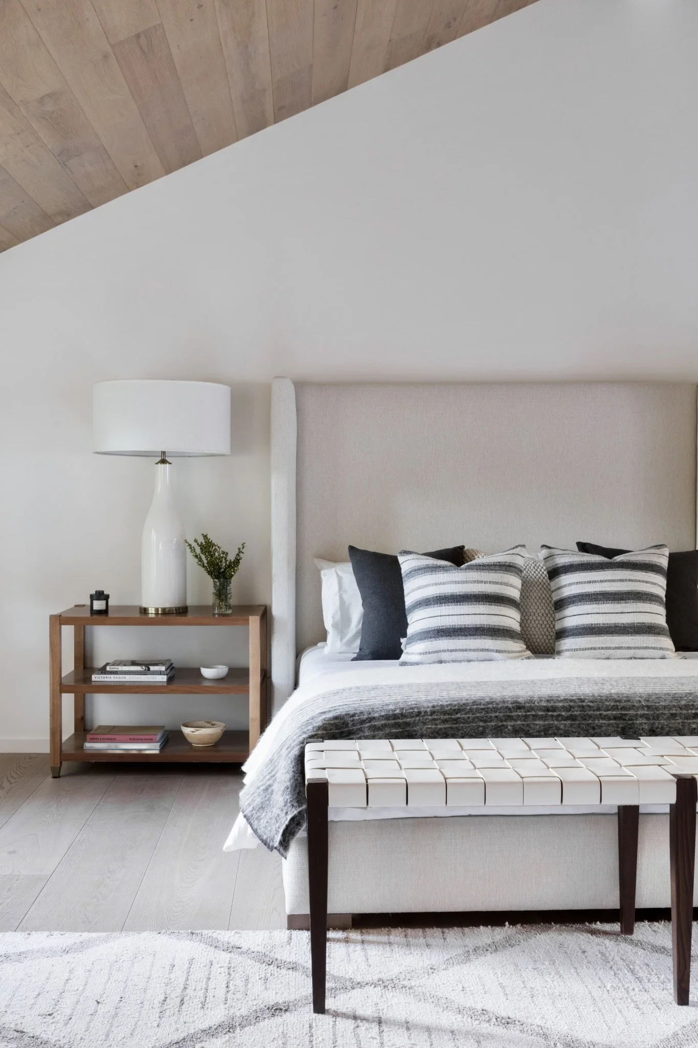 A bedroom with a beige bed, striped and solid pillows, a dark blanket, a white bench at the foot of the bed, a wooden nightstand with books, a small plant, a lamp, and a small bowl, Rachael Goddard Design Studio Calabasas