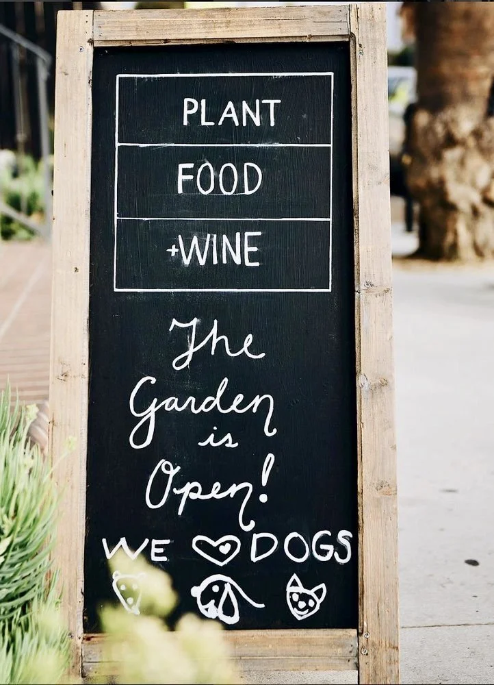 Rachael Goddard Design StudioOutdoor chalkboard sign with a wooden frame advertising plant, food, and wine, and indicating the garden is open. Handwritten text reads 'The garden is open! We ❤️ dogs' with doodles of a dog and a dog face at the bottom.