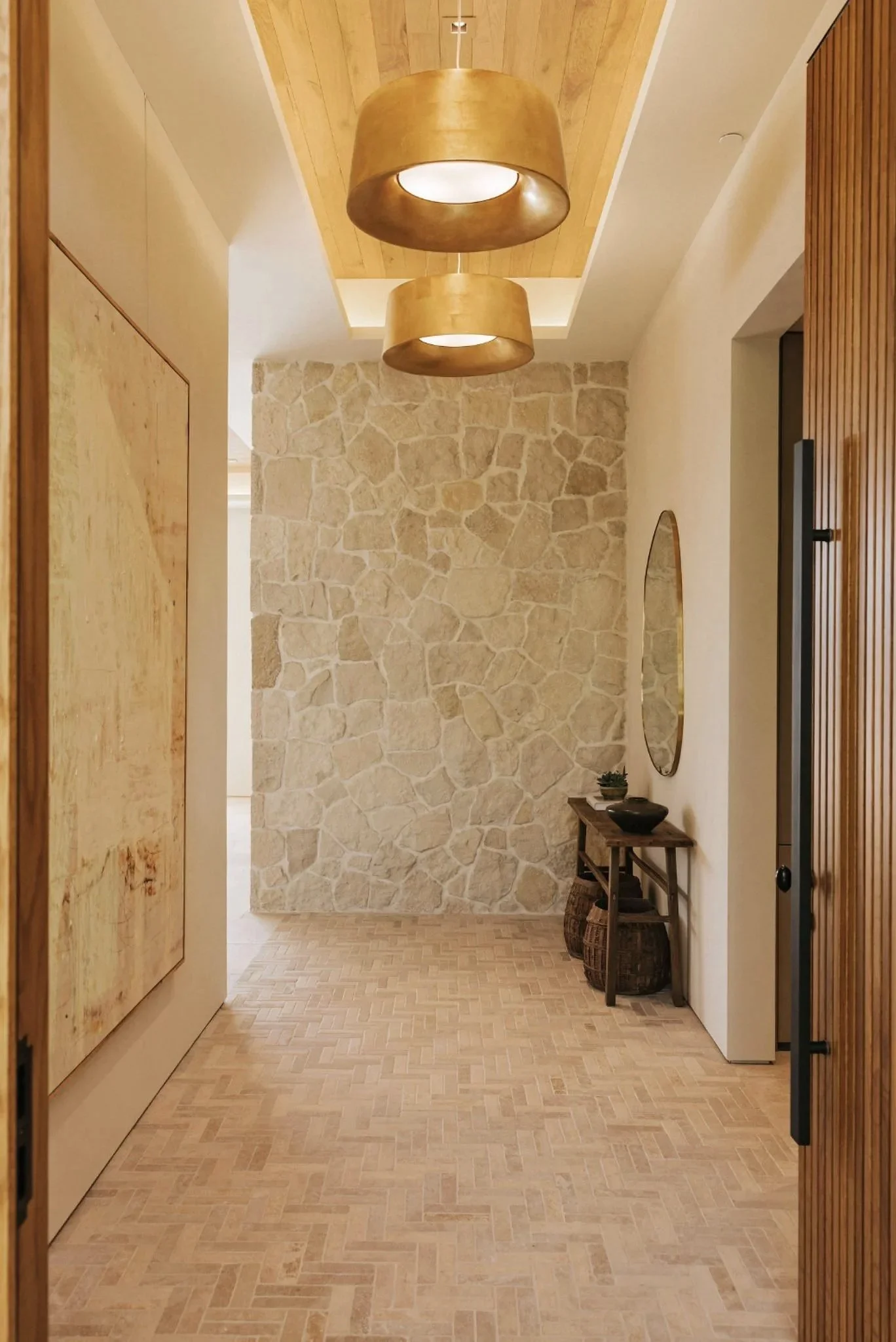 Rachael Goddard Design Studio Calabasas Entryway with wooden flooring, a stone accent wall, a small wooden table with two baskets underneath, a mirror on the wall, and two large, gold-colored pendant lights hanging from the ceiling.