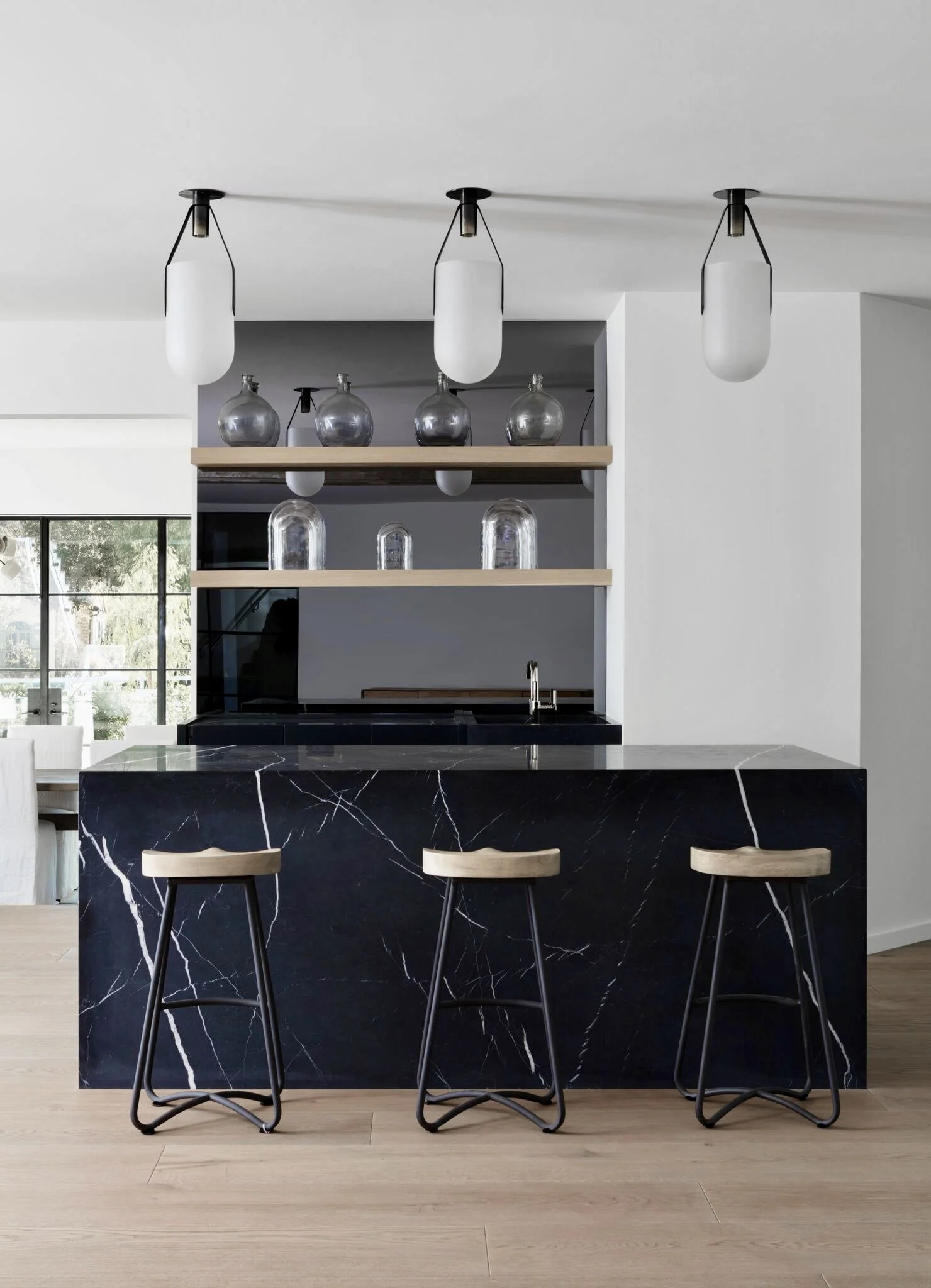 Modern kitchen with a black marble island, three bar stools, open shelves with decorative glass bottles, and hanging pendant lights.  Rachael Goddard Design Studio Calabasas