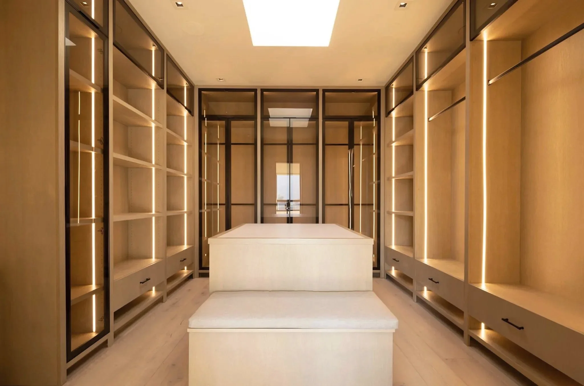 Rachael Goddard Design Studio Calabasas Empty walk-in closet with built-in shelves, glass doors, and an island in the center, illuminated by ceiling lighting.