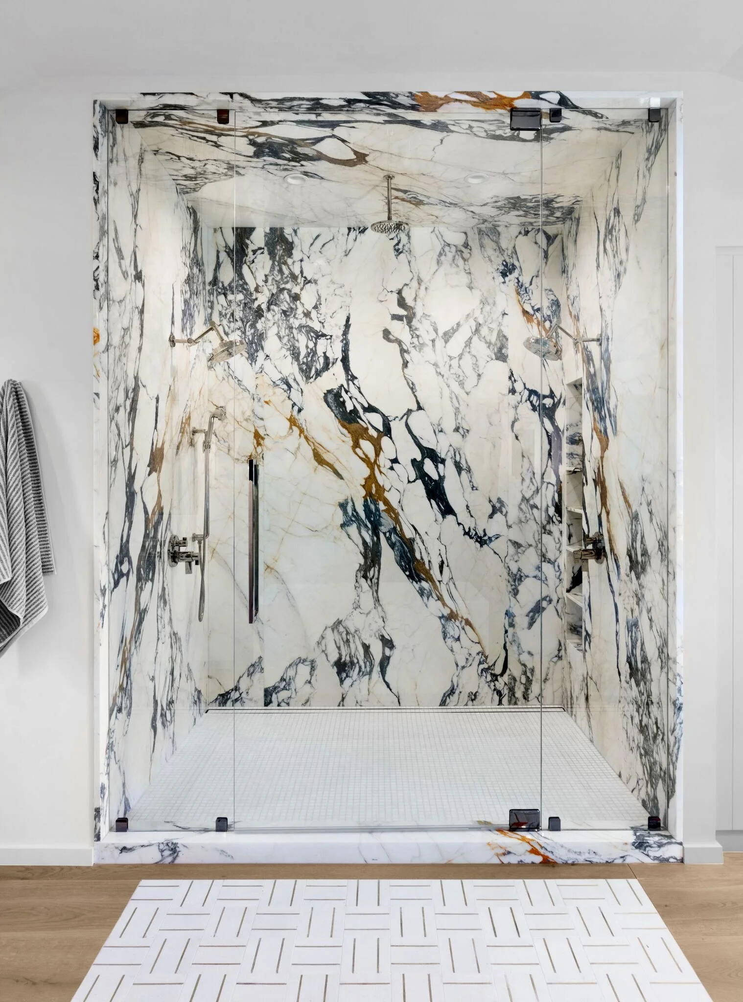 Modern shower with marble walls and glass doors, featuring multiple showerheads and built-in shelves.  Rachael Goddard Design Studio Calabasas