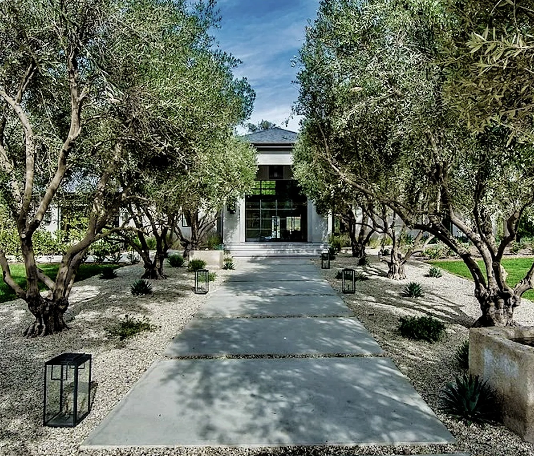A modern house at the end of a concrete pathway lined with trees and outdoor lanterns.  Rachael Goddard Design Studio Calabasas