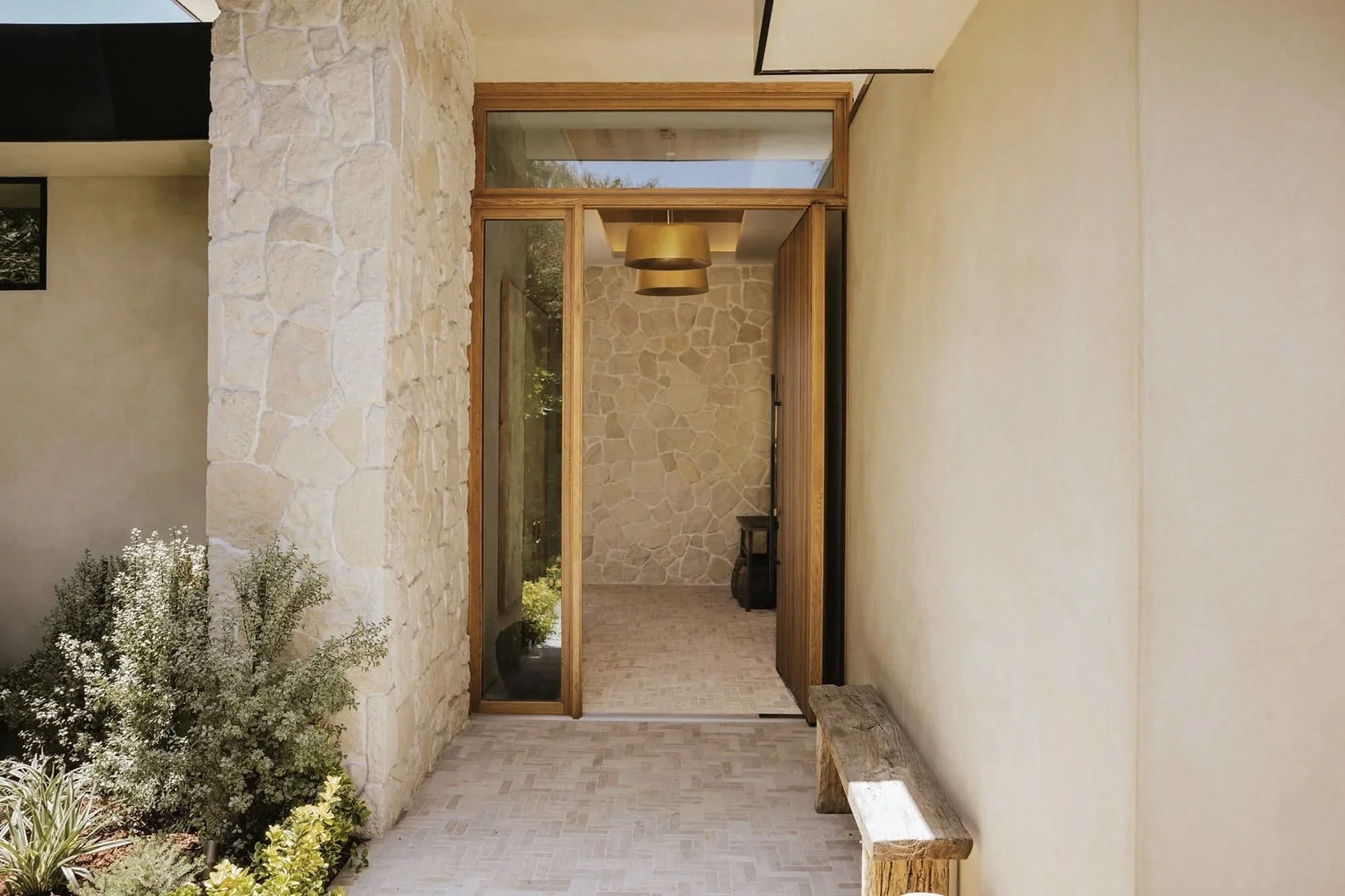 Rachael Goddard Design Studio Calabasas Entrance area of a modern house with stone and wooden accents, a bench, and some greenery outside.