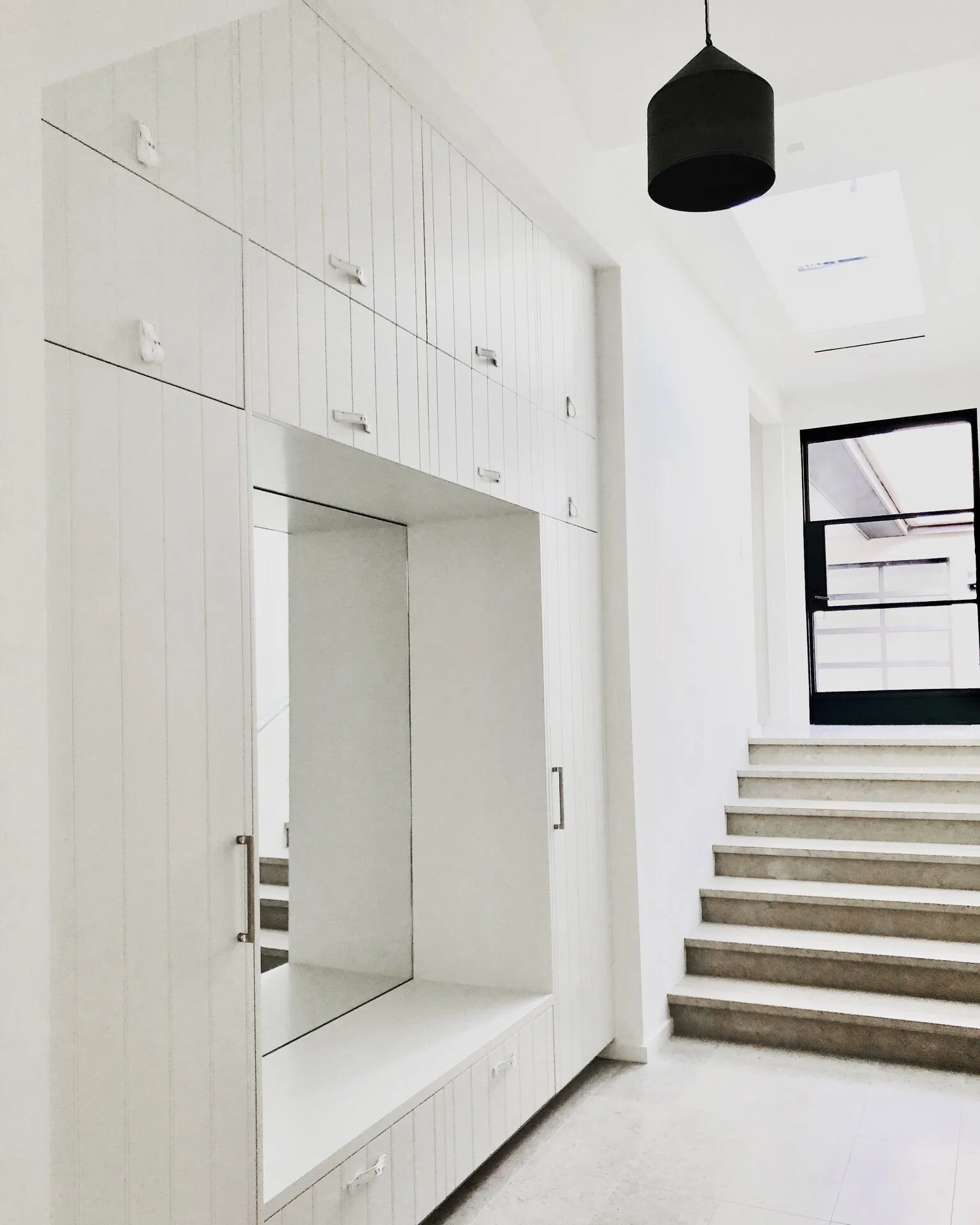 Modern interior space with white built-in cabinets, a large mirror, and a staircase with beige steps leading to a black-framed glass door, natural light from skylights, and a black pendant ceiling light.  Rachael Goddard Design Studio Calabasas
