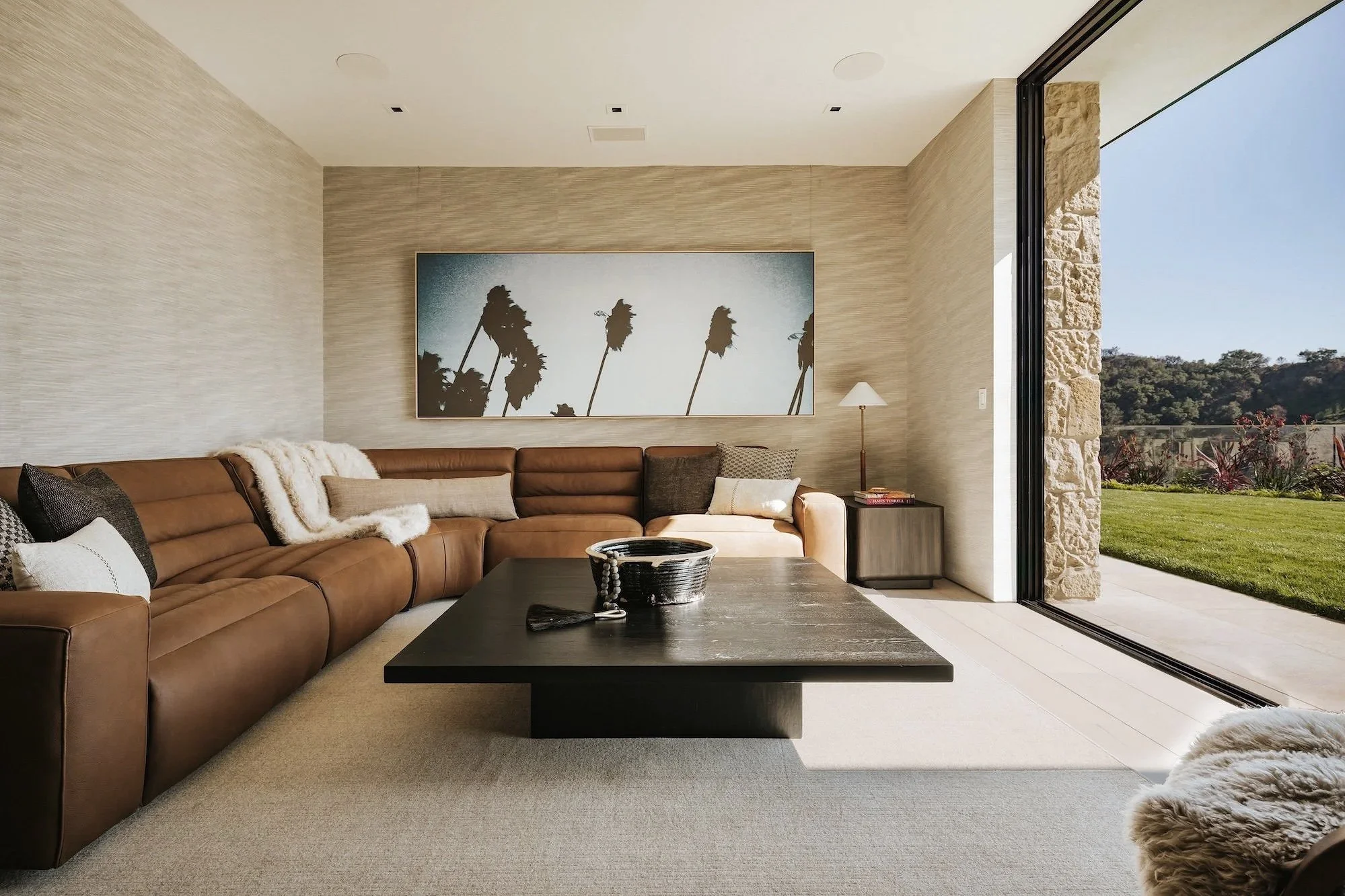 Rachael Goddard Design Studio Calabasas Modern living room with brown leather sectional sofa, large wall art of palm trees, and an open sliding door leading outside to a grassy yard with trees.