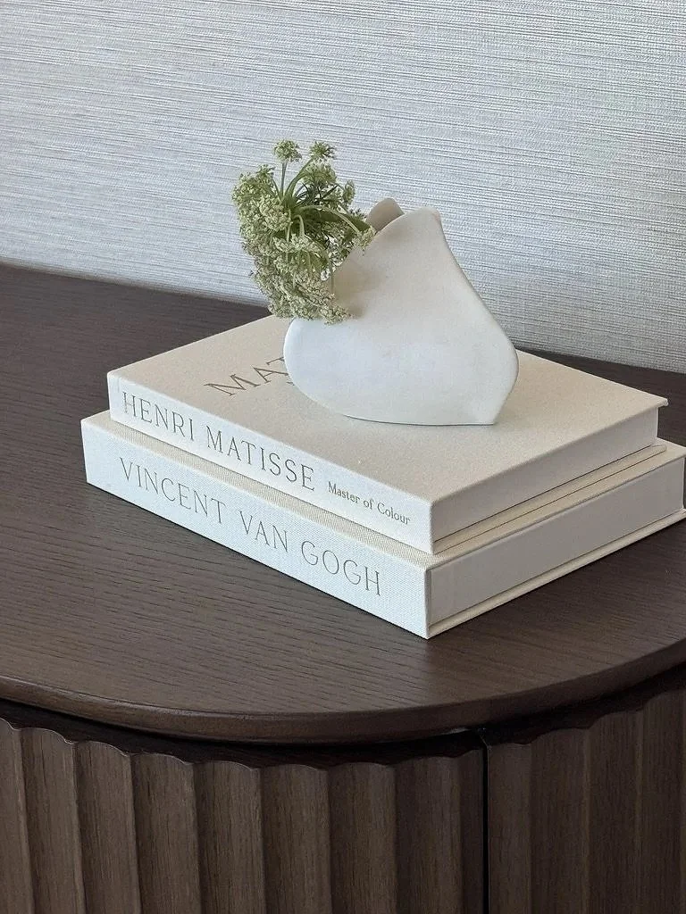 Rachael Goddard Design Studio Calabasas A white vase with small white flowers sits atop two large art books titled "Henri Matisse Master of Colour" and "Vincent Van Gogh" on a dark wooden table against a light textured wall.