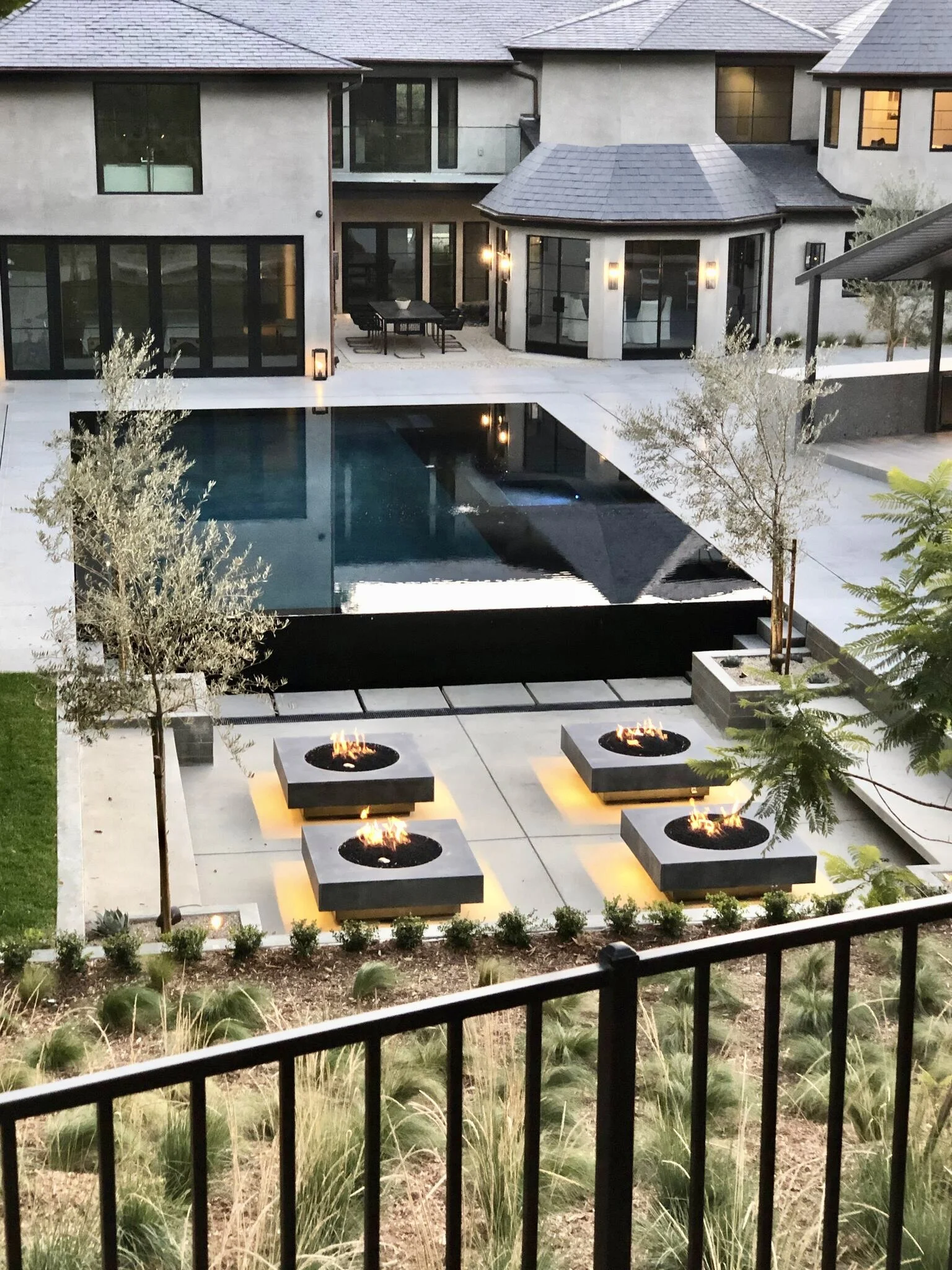 Modern backyard view of a house with a black swimming pool, outdoor fire pits on concrete patios, trees, and outdoor seating area, with large windows and a balcony.  Rachael Goddard Design Studio Calabasas