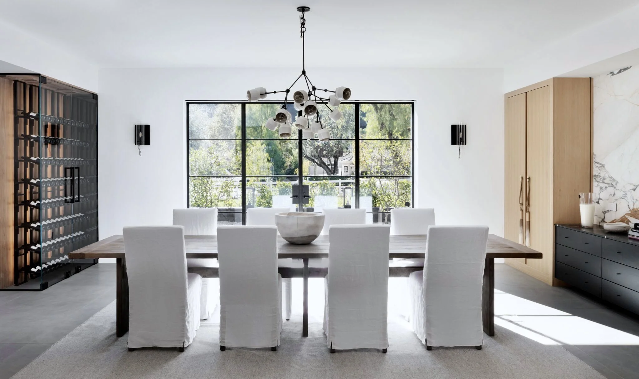 Modern dining room with a large wooden table, eight white slipcovered chairs, a black sideboard, a wine fridge, a chandelier, and a large window with greenery outside.  Rachael Goddard Design Studio Calabasas