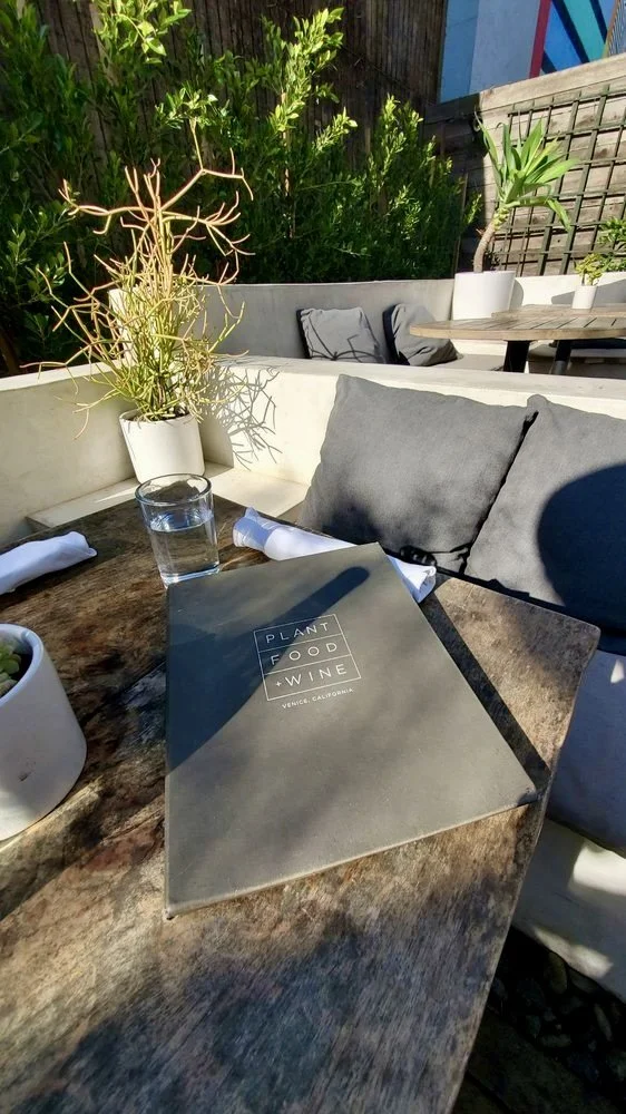 Rachael Goddard Design Studio Calabasas Outdoor restaurant table with a menu labeled 'Plant Food Wine' from Venice, California, a glass of water, and potted plants, surrounded by chairs and greenery.