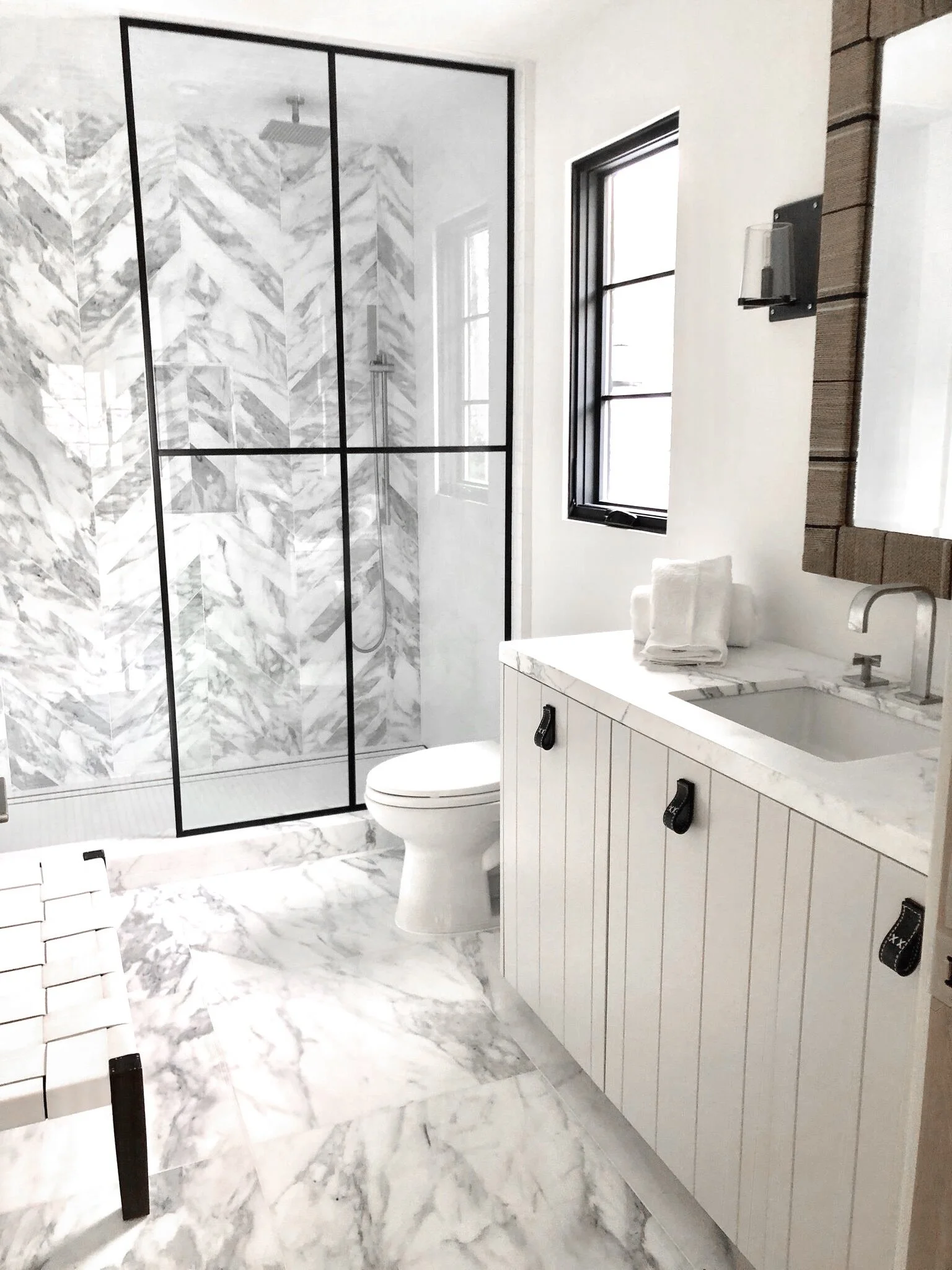 A modern bathroom with a white marble floor and wall tiles, a walk-in shower with a glass enclosure, a white toilet, a vanity with a marble countertop, a small sink, and a black-framed window.  Rachael Goddard Design Studio Calabasas