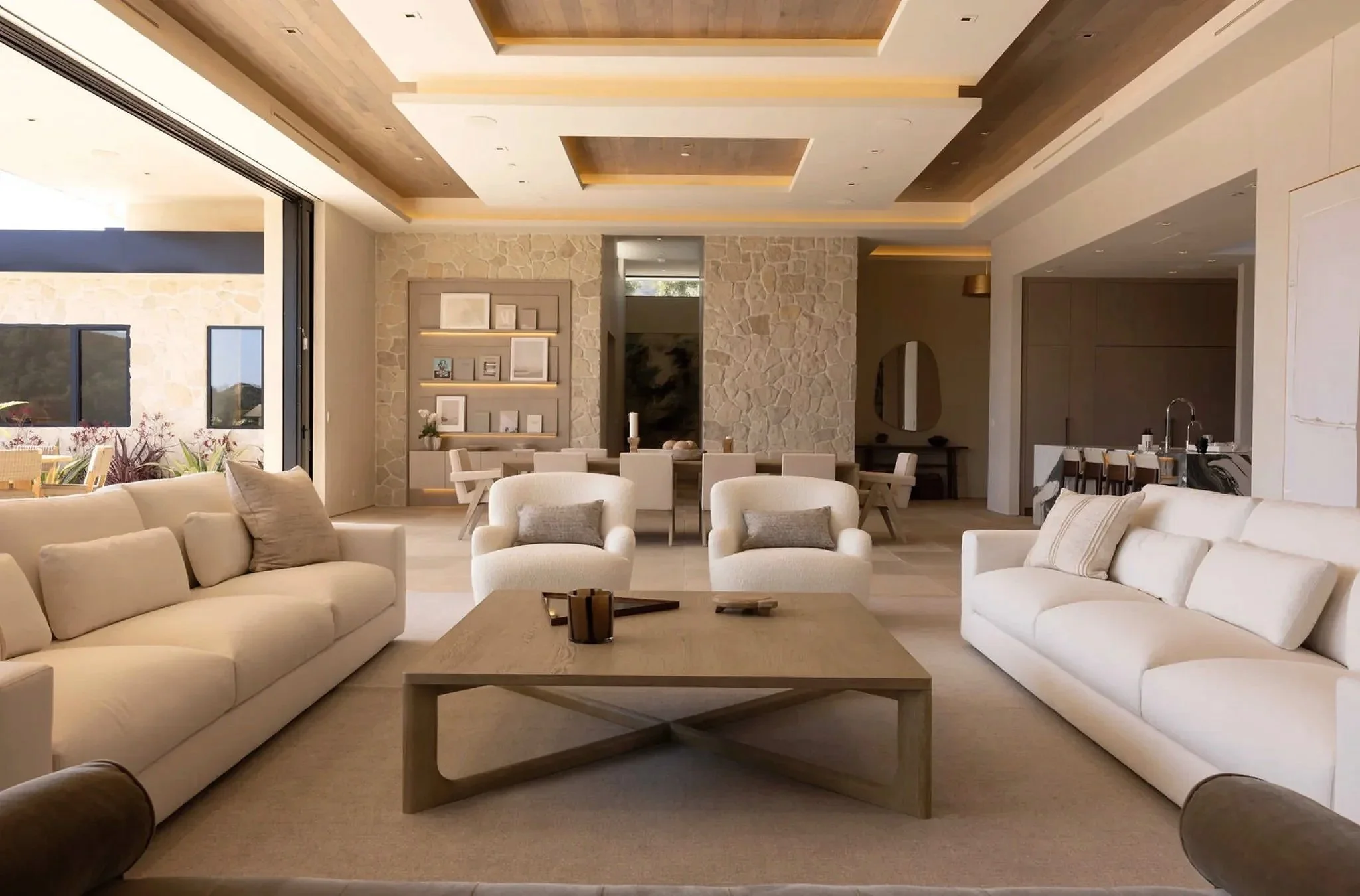 Rachael Goddard Design Studio Calabasas Modern living room with beige sofas, armchairs, wooden coffee table, and a stone accent wall with built-in shelving containing picture frames. Open floor plan with dining area, large windows, and indoor plants.