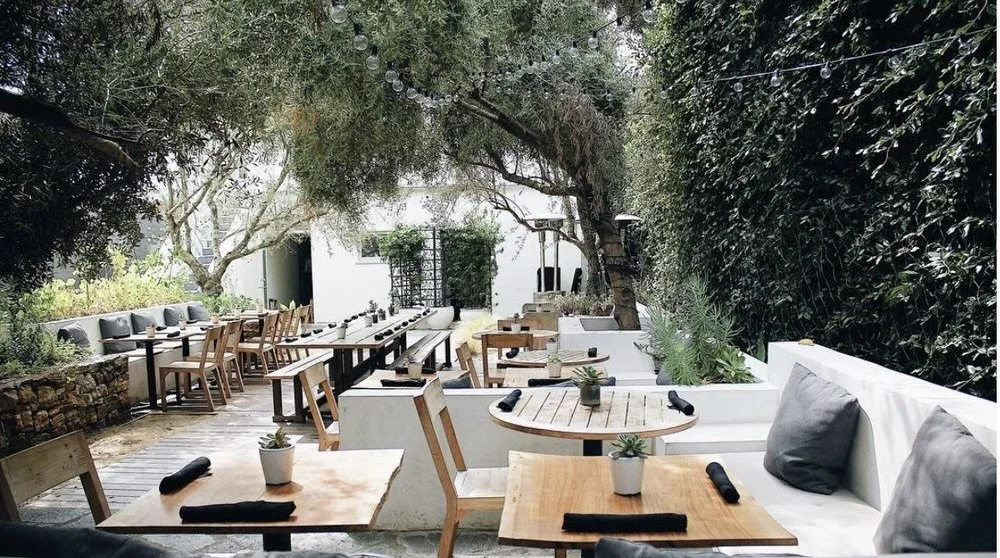 Rachael Goddard Design Studio Calabasas Outdoor restaurant with wooden tables, chairs, and built-in white seating, decorated with small plants, under string lights, with lush trees and a hedge background.