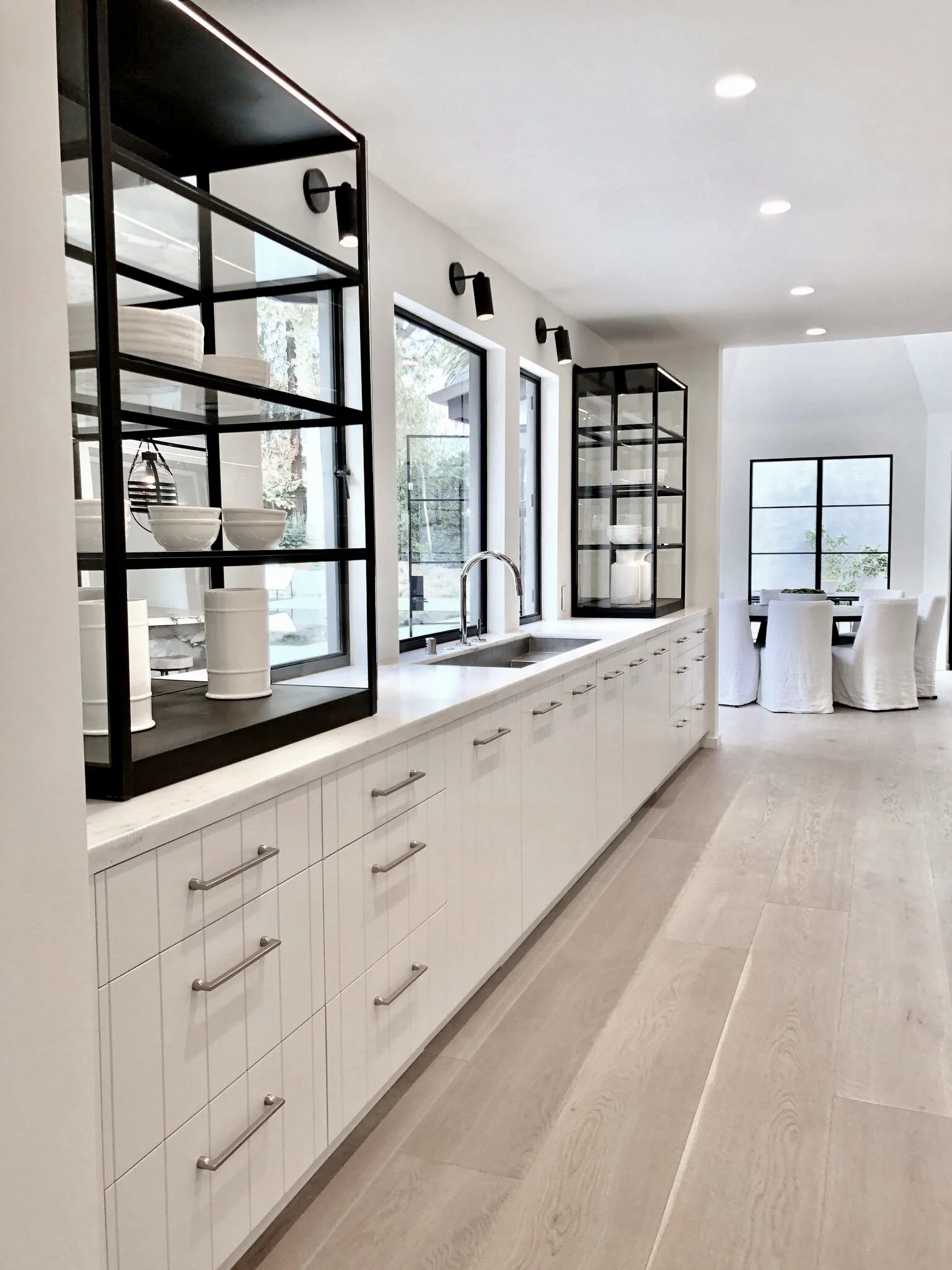 Modern kitchen with white cabinetry, black open shelves, large windows, a sink with a faucet, and a dining area with white chairs in the background.  Rachael Goddard Design Studio Calabasas