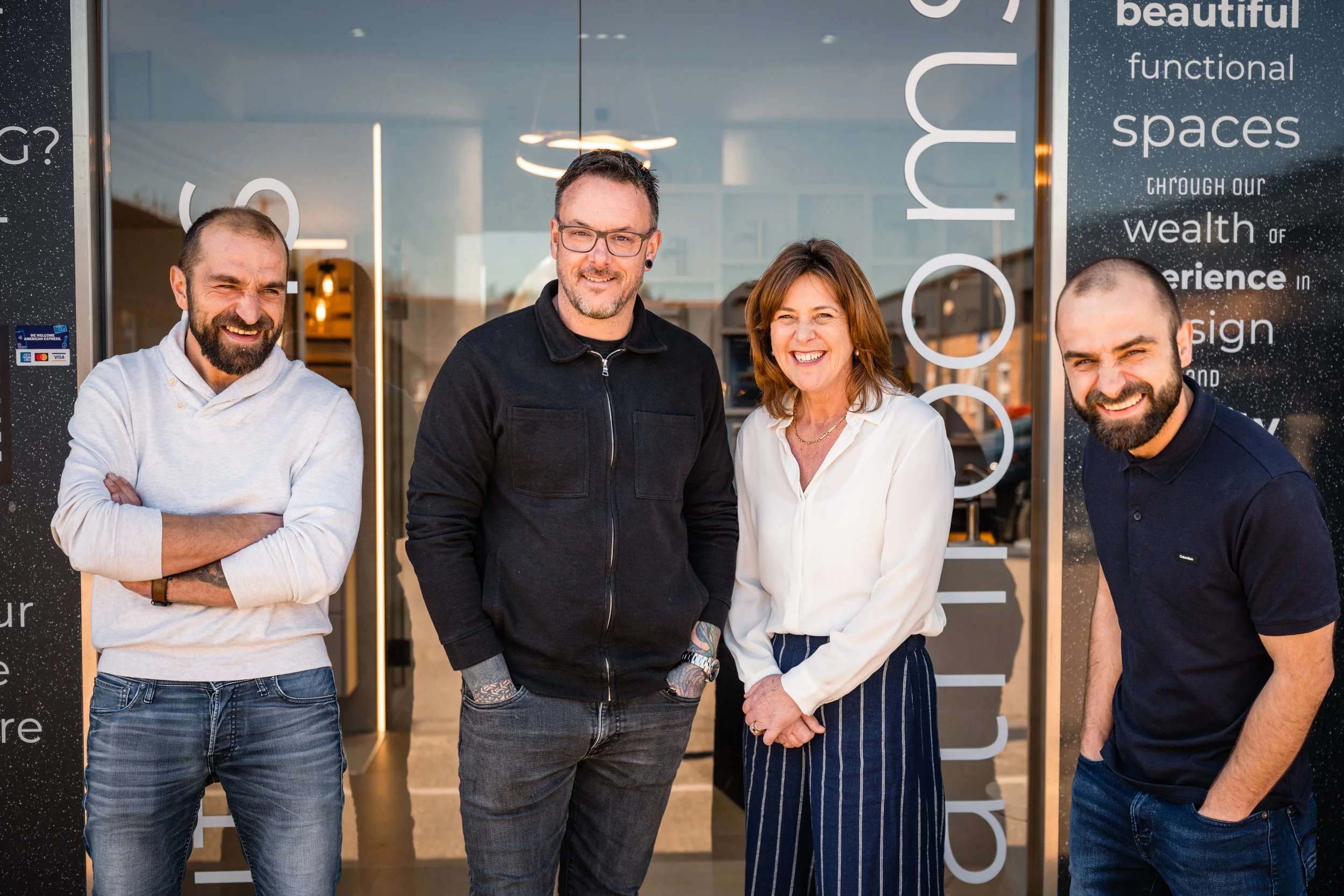 Group of four smiling people standing outside a modern business or office building with glass doors and windows.