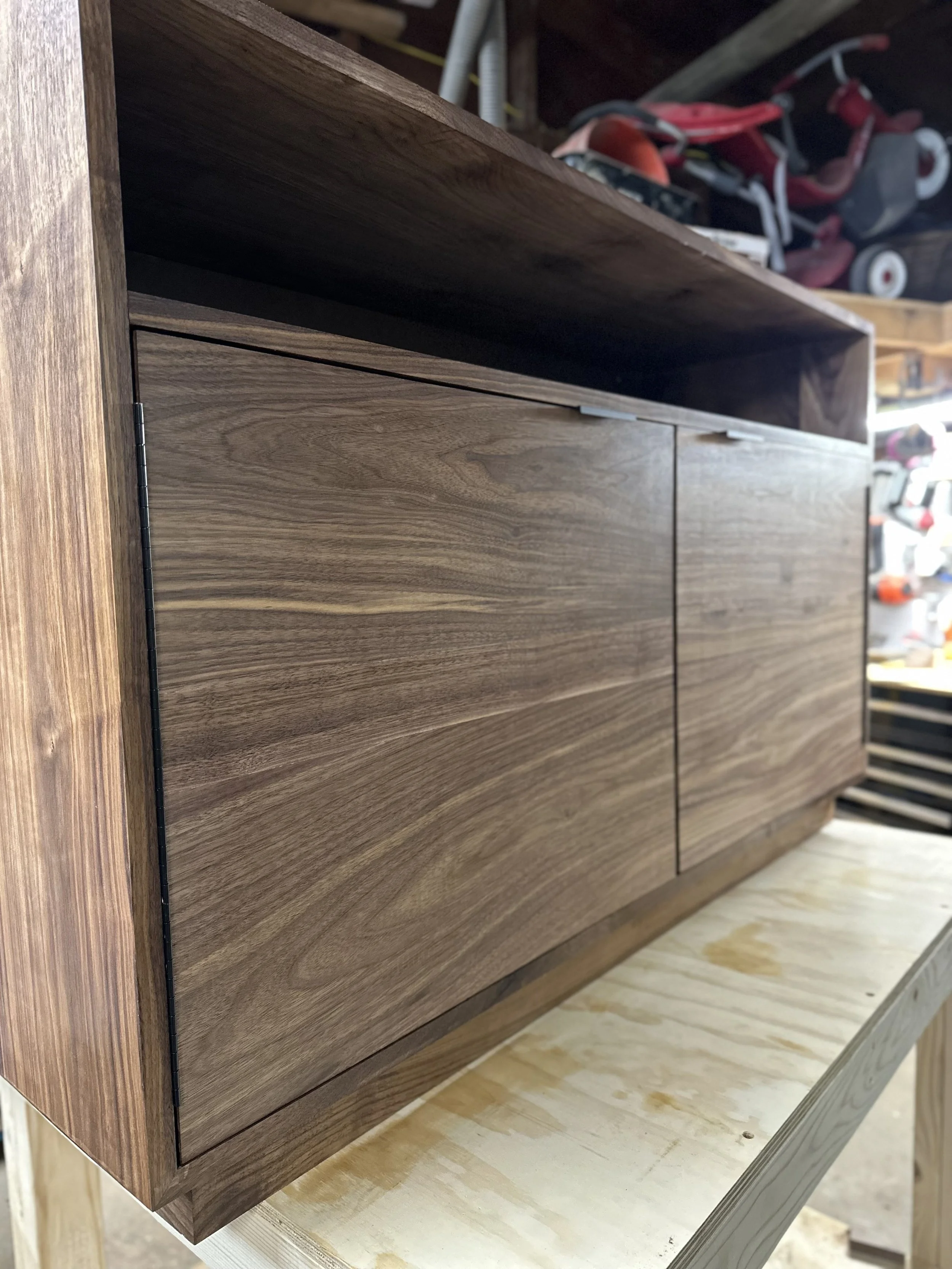 Close-up of a wooden cabinet with a natural finish, placed on a light-colored wooden workbench in a woodworking shop, with tools visible in the background.