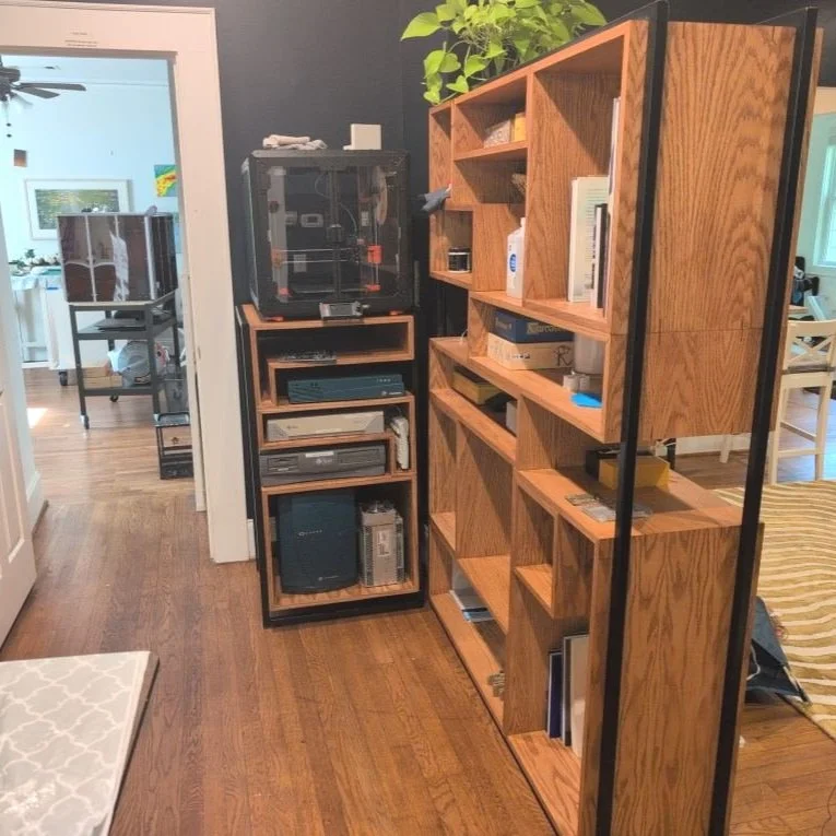 Wooden shelving unit with books and various objects, adjacent to a black plastic storage cabinet, in a hallway with hardwood flooring and an open door leading to a dining area.