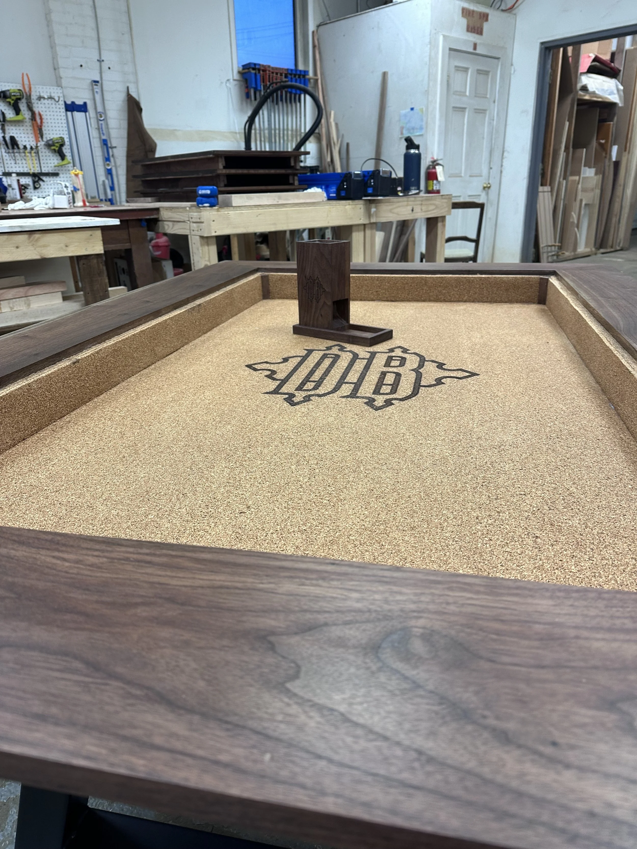 A wooden table with sand and a monogrammed logo, in a woodworking workshop with tools, wood, and supplies in the background.