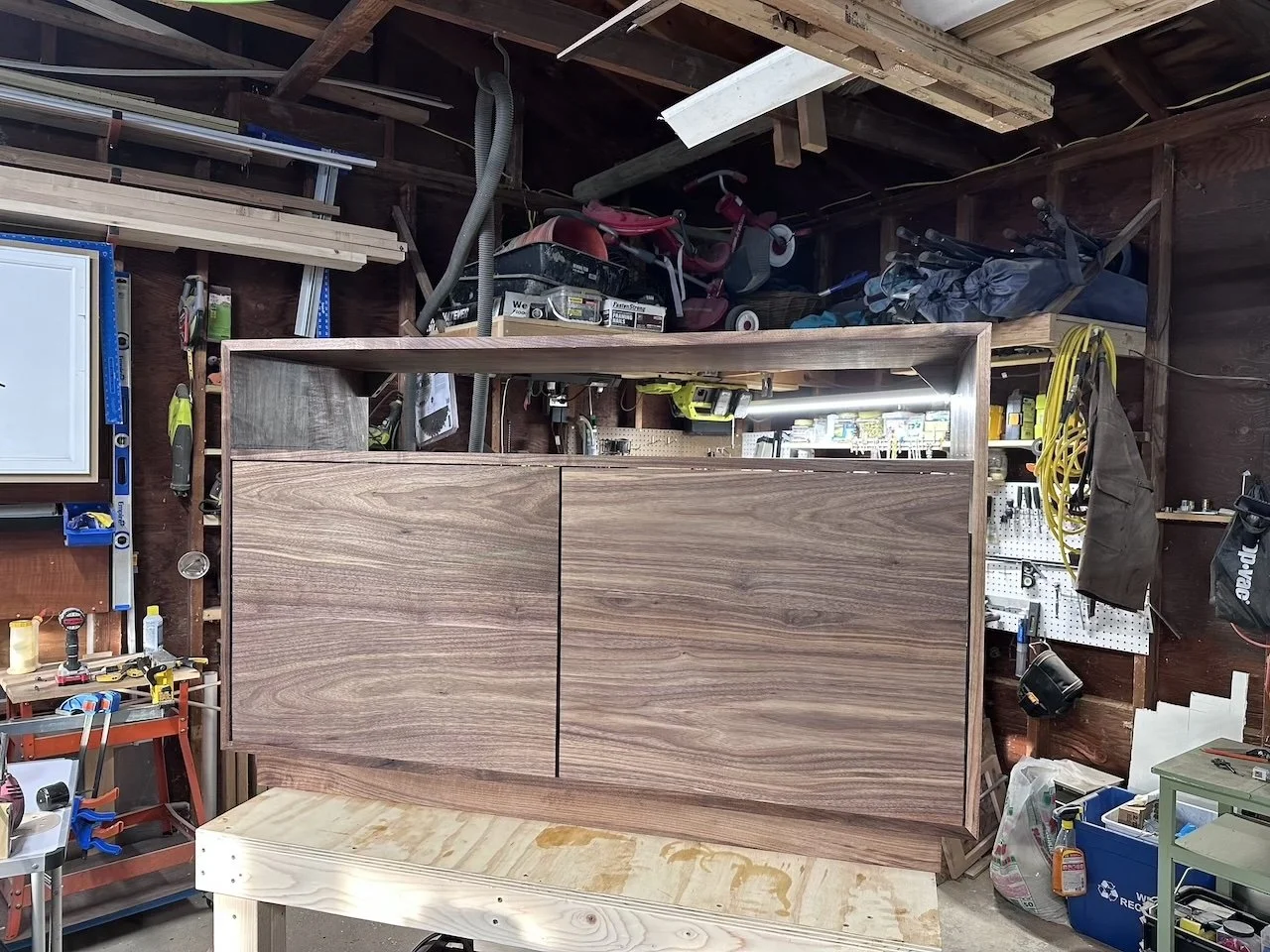A woodworking project in progress in a cluttered garage, featuring a partially assembled wooden cabinet with a natural wood finish, surrounded by tools and storage items.